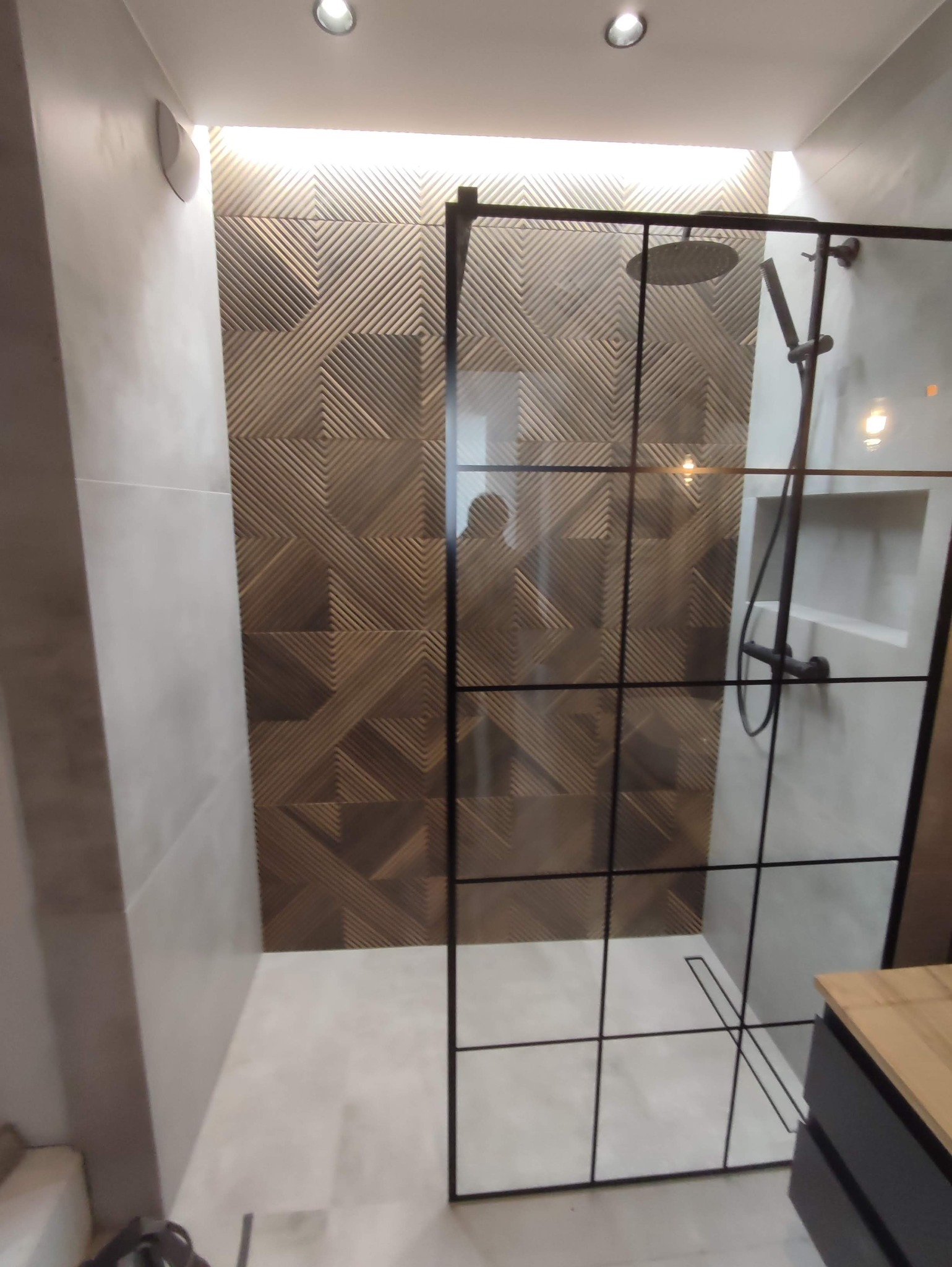 Nowoczesna kabina prysznicowa z czarnymi profilami i deszczownicą, ściana wykończona dekoracyjnymi panelami z geometrycznym wzorem w odcieniach brązu i beżu, minimalistyczna aranżacja łazienki.