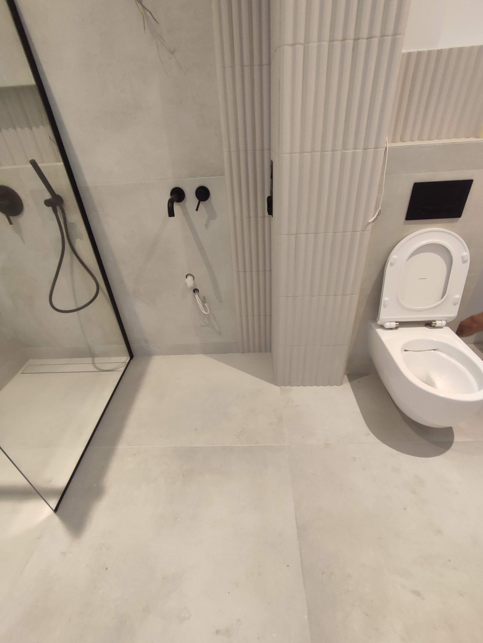 Nowoczesna łazienka w odcieniach szarości z czarną armaturą, kabiną prysznicową typu walk-in i podwieszaną toaletą z deską w górze, wykończona płytkami imitującymi beton i dekoracyjnymi panelami...