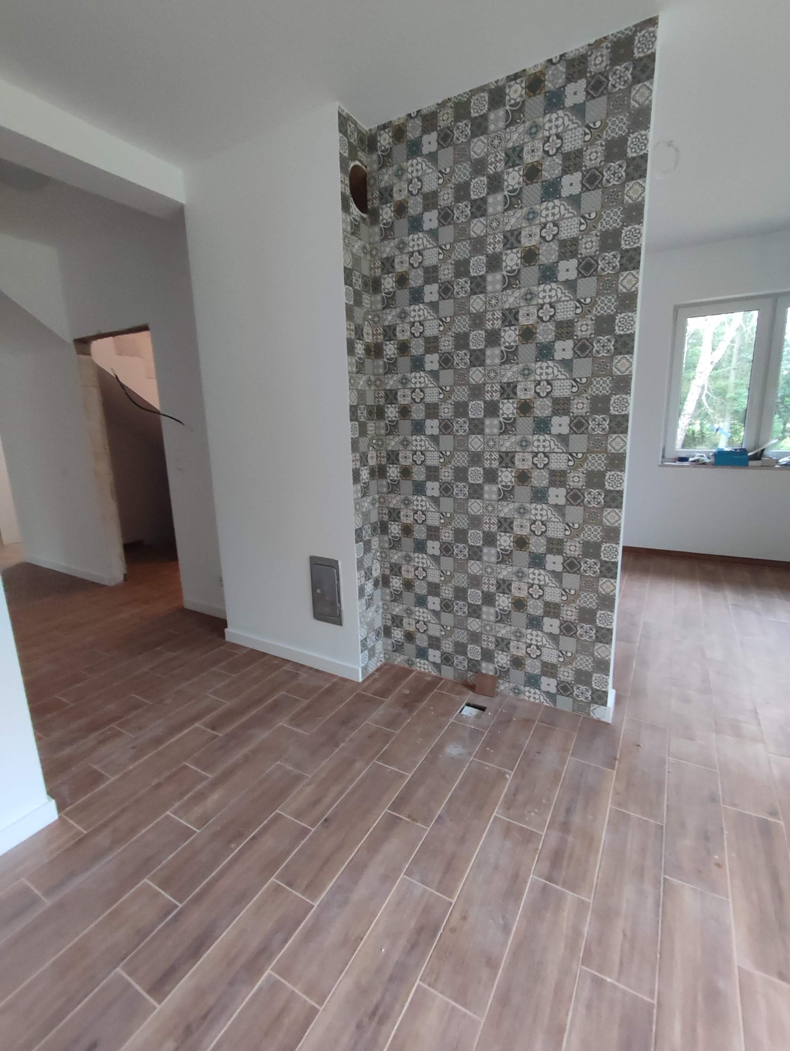 Ukończone układanie podłogi z płytek imitujących drewno w nowym pomieszczeniu, z fragmentem ściany wykończonej dekoracyjnymi płytkami w stylu patchwork, widoczny otwór w podłodze.
