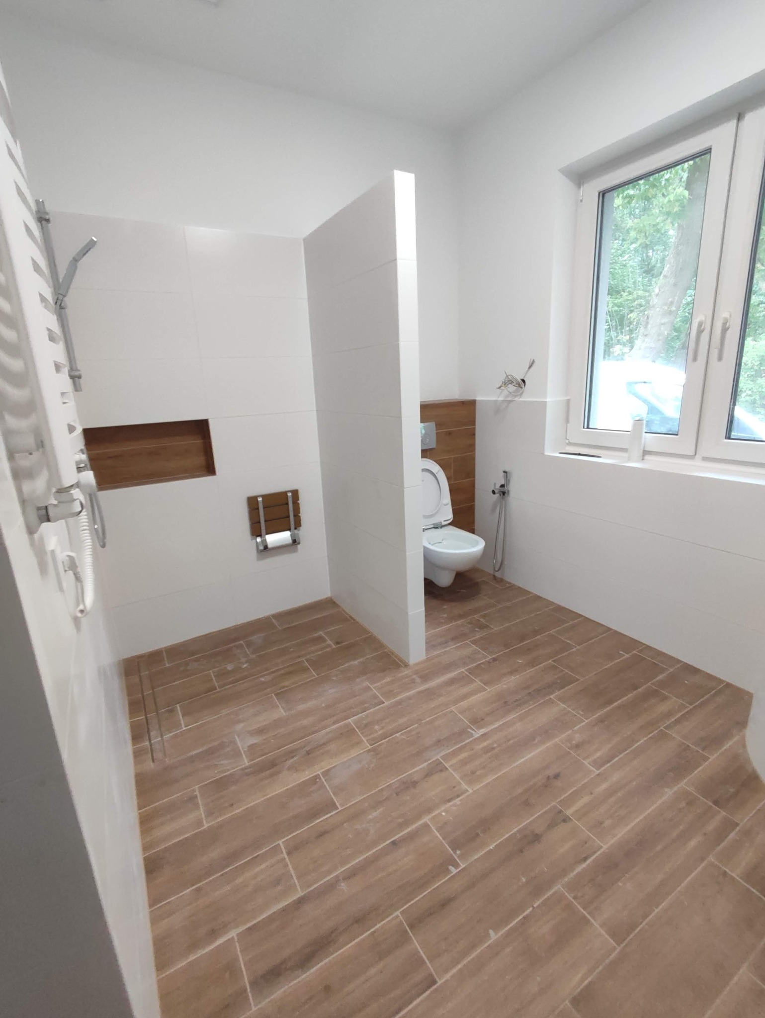 Nowoczesna łazienka z brązowymi płytkami podłogowymi imitującymi drewno, białe płytki ścienne, kabina prysznicowa typu walk-in oraz toaleta z bidetką, widok z okna na zieleń.