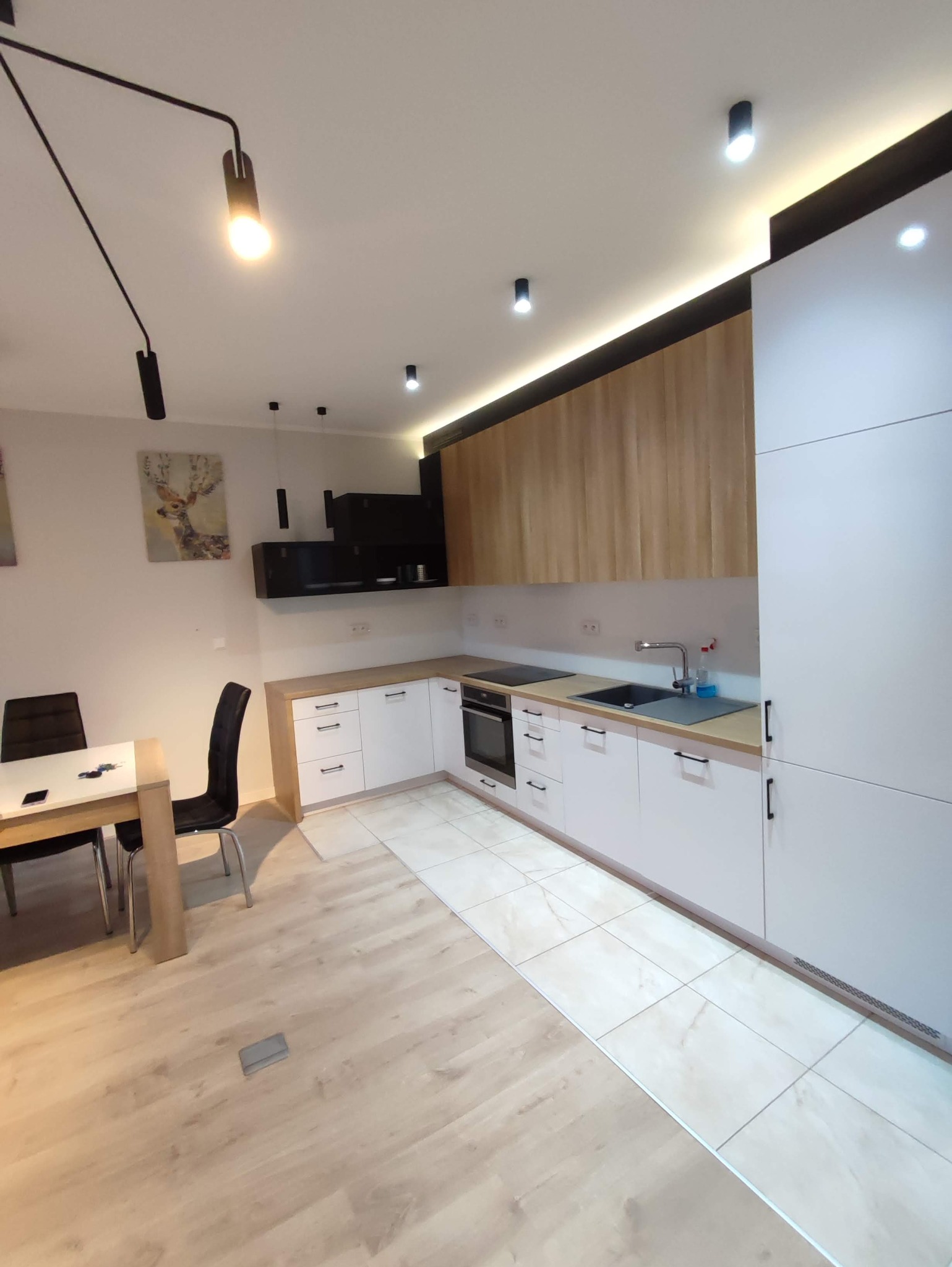 Nowoczesna kuchnia z białymi szafkami, drewnianymi akcentami i oświetleniem punktowym; widoczny stół z krzesłami i obraz z motywem jelenia na ścianie.