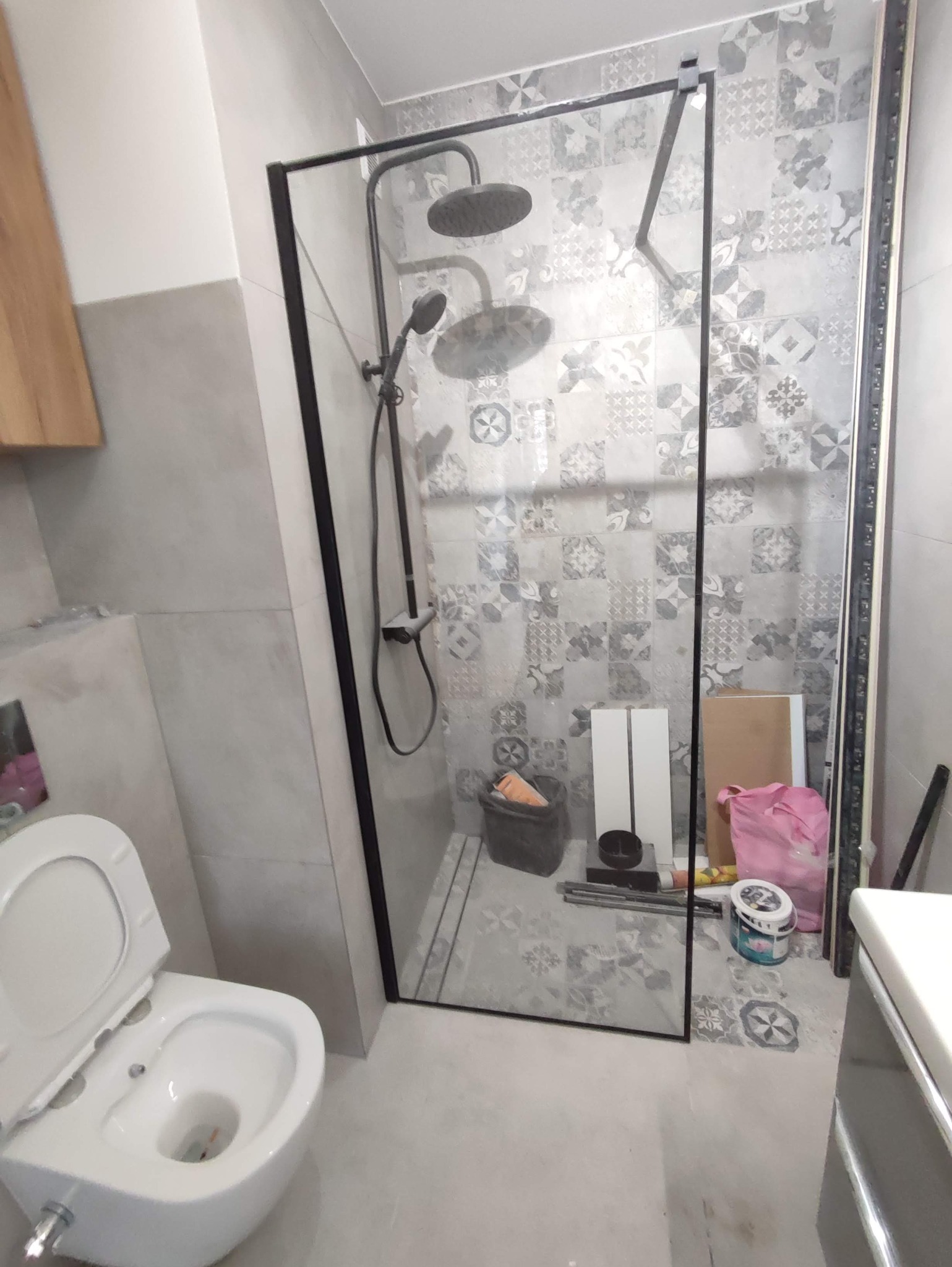 Łazienka w trakcie prac wykończeniowych: zamontowana kabina prysznicowa z czarną ramą i deszczownicą, widoczna toaleta, ściany wyłożone płytkami w patchworkowy wzór, narzędzia i materiały budowlane...