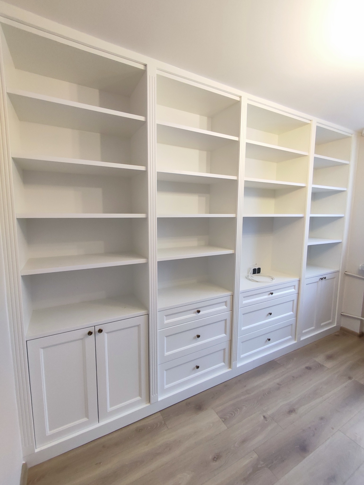 Biała zabudowa meblowa z szafkami i szufladami, tworząca biblioteczkę z pustymi półkami, z delikatnymi zdobieniami frezowanymi na obrzeżach.