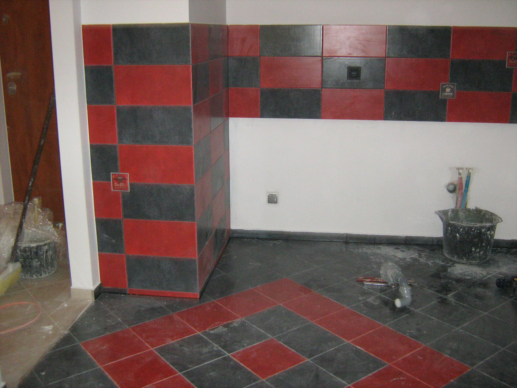 Układanie płytek podłogowych i ściennych w szachownicę z czerwono-czarnych płytek w trakcie prac wykończeniowych, widoczne narzędzia i wiadro.