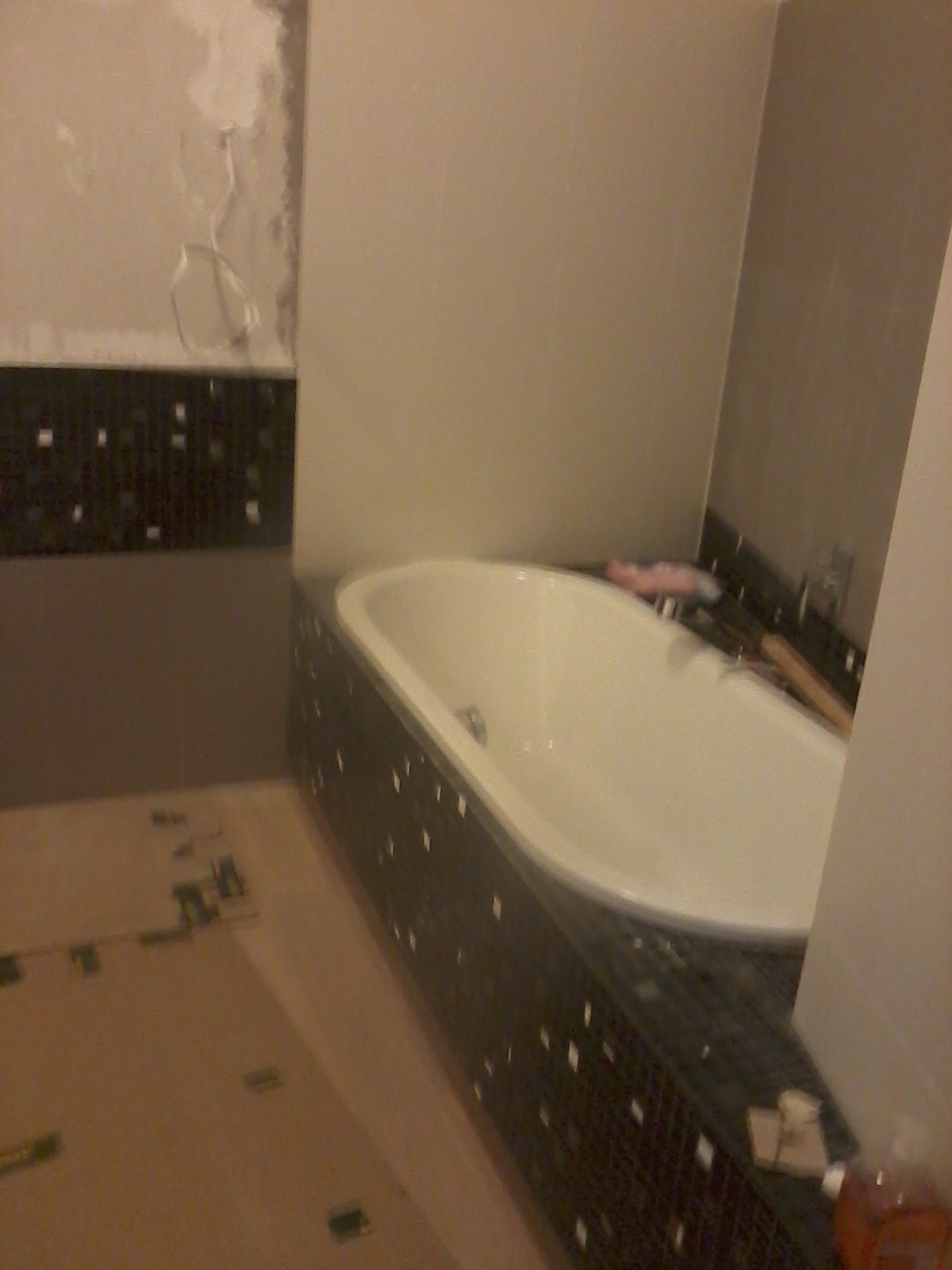 Biała wanna obudowana mozaiką w trakcie montażu, widoczne niedokończone fragmenty ściany i zabezpieczona podłoga.