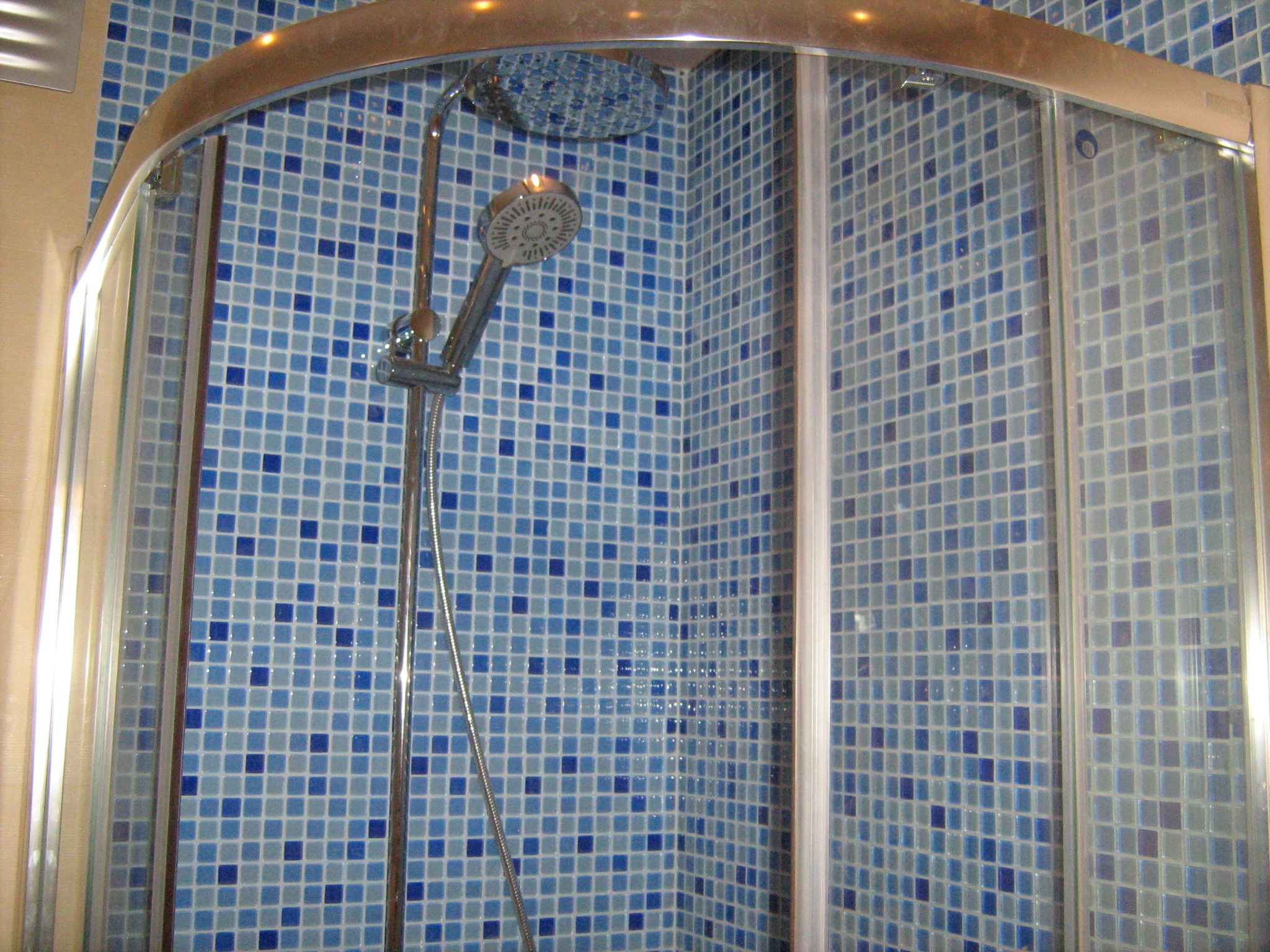 Kabina prysznicowa z mozaiką w odcieniach niebieskiego, chromowana armatura, oświetlenie sufitowe odbijające się w powierzchni kabiny.