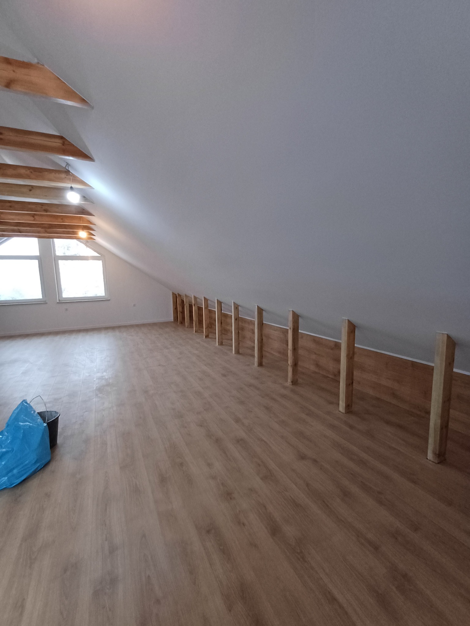 Poddasze z drewnianą podłogą i przygotowaną konstrukcją z drewnianych słupków przy ścianie skośnej, widoczne okna dachowe i worki na śmieci.