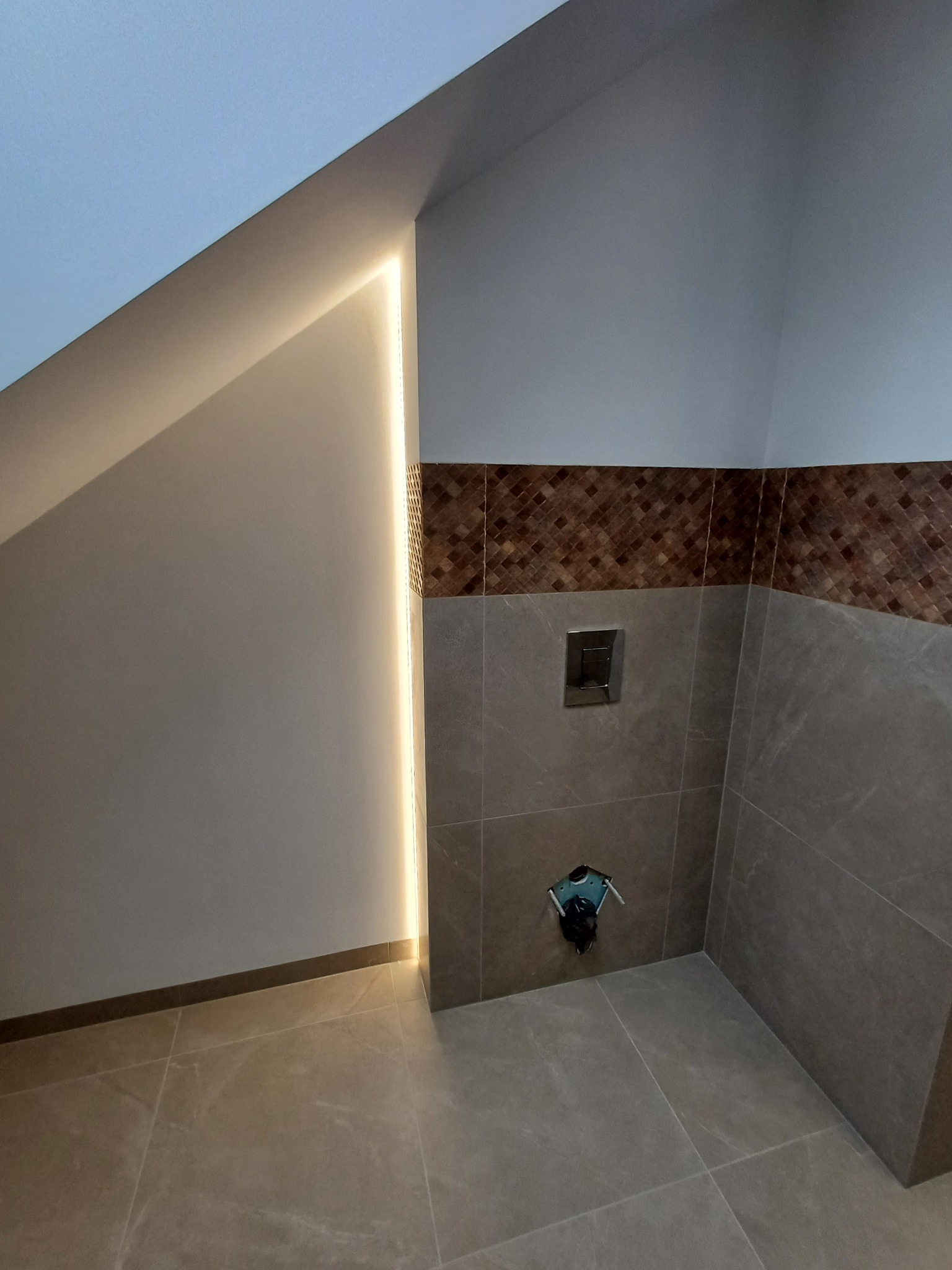 Nowoczesna łazienka na poddaszu z ukośną ścianą i oświetleniem LED wzdłuż krawędzi, brązowa mozaika nad szarymi płytkami, widoczny stelaż podtynkowy.