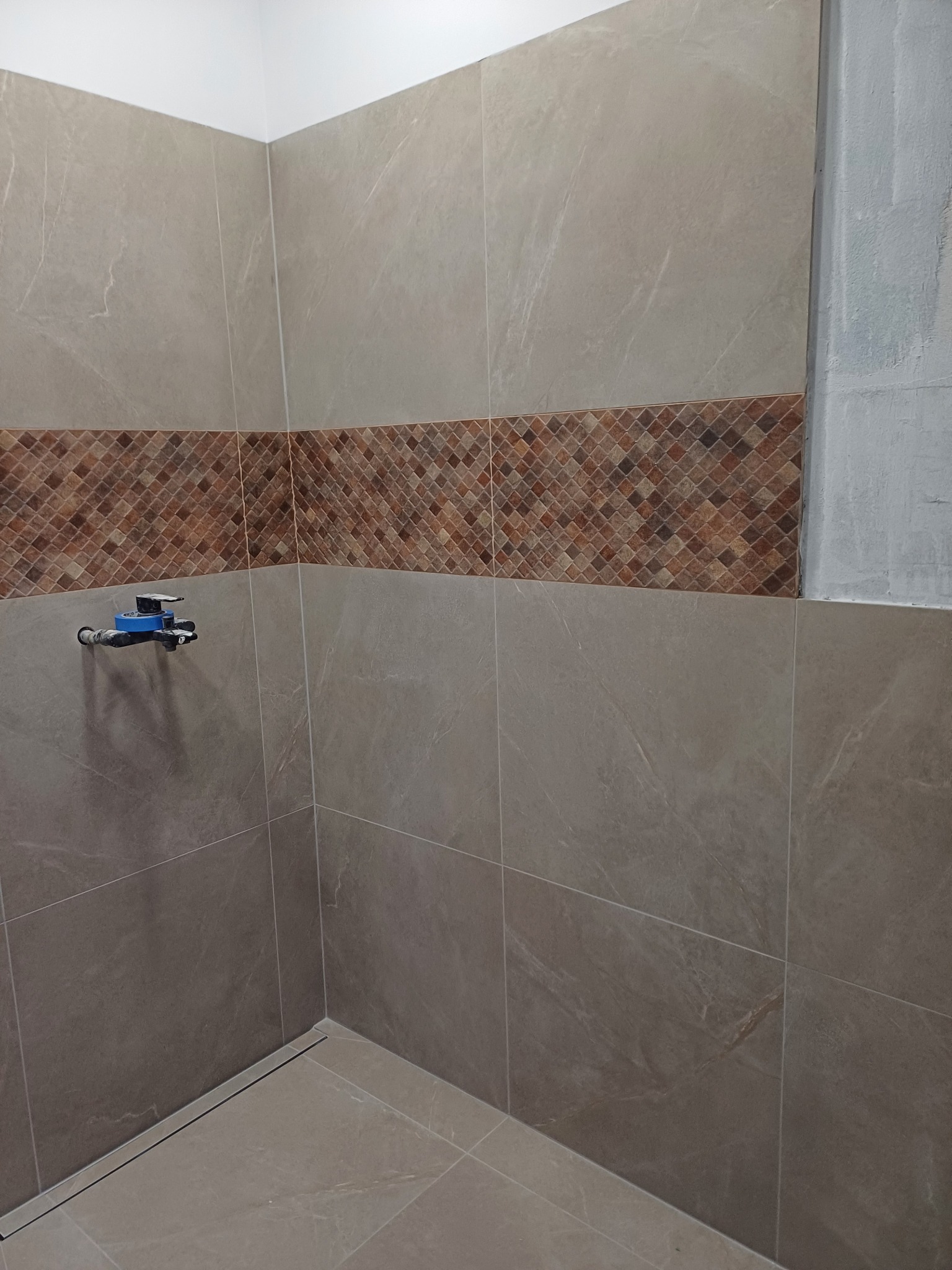 Fragment łazienki w trakcie remontu: ułożone płytki ścienne w odcieniach beżu z dekoracyjnym pasem brązowej mozaiki, widoczna instalacja wodna zabezpieczona niebieską taśmą, ślad po starej glazurze...