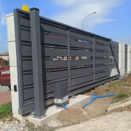 Nowoczesna, przesuwna brama w kolorze grafitowym z betonowymi słupkami, widoczny napęd bramy i niebieskie rury instalacyjne w ziemi.