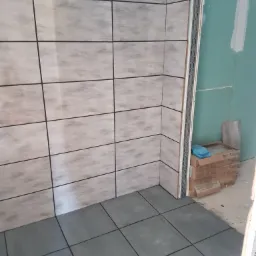 Fragment łazienki w trakcie wykańczania: ułożone szare płytki podłogowe i beżowe płytki ścienne z ciemną fugą, widoczne profile narożne i kartony z zapasem płytek przy ścianie z płyt...