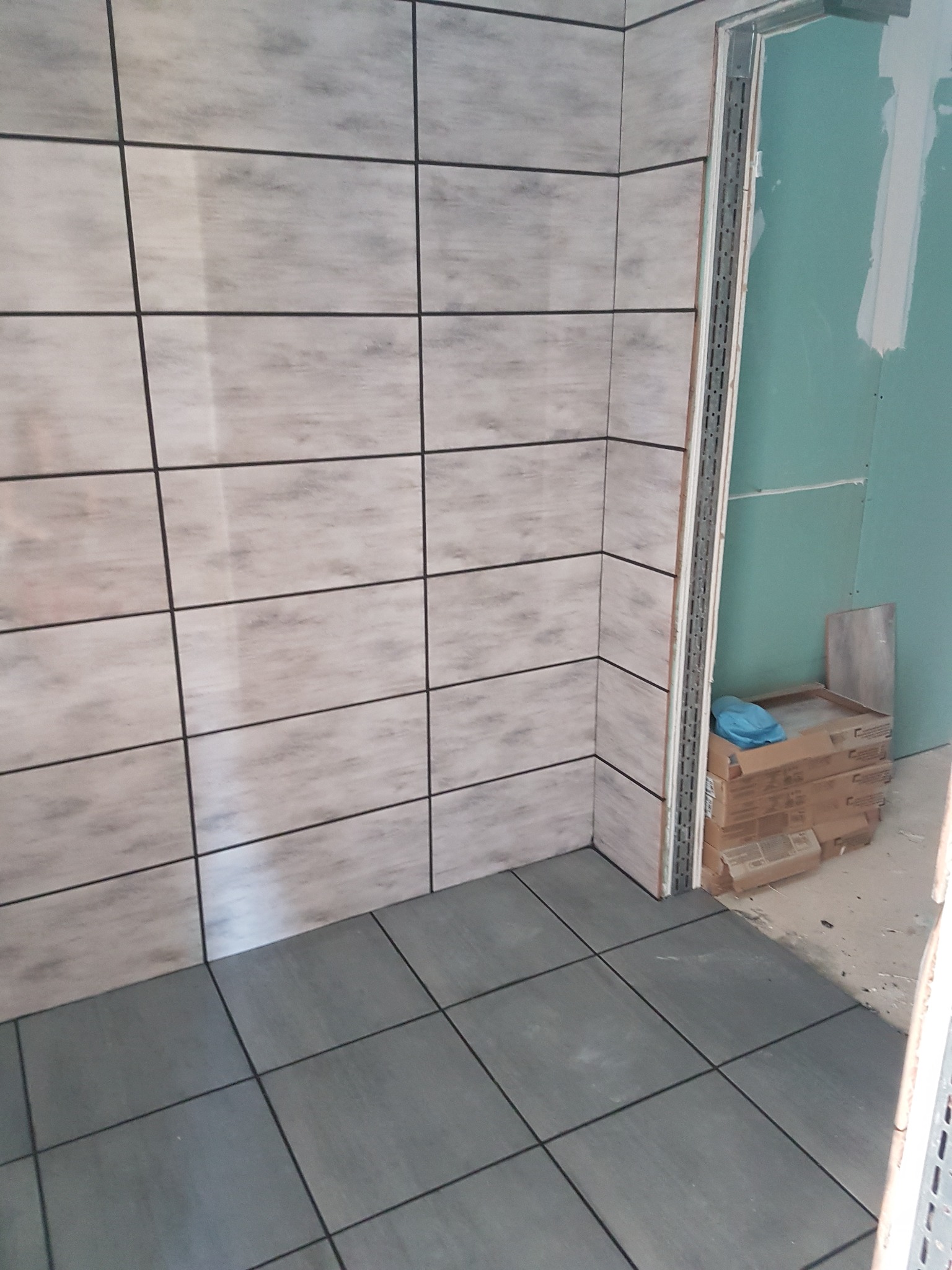 Fragment łazienki w trakcie wykańczania: ułożone szare płytki podłogowe i beżowe płytki ścienne z ciemną fugą, widoczne profile narożne i kartony z zapasem płytek przy ścianie z płyt...