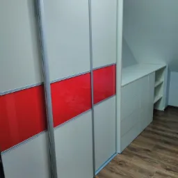 Biała szafa przesuwna z czerwonymi akcentami w zabudowie pod skosem, z drewnianą podłogą.