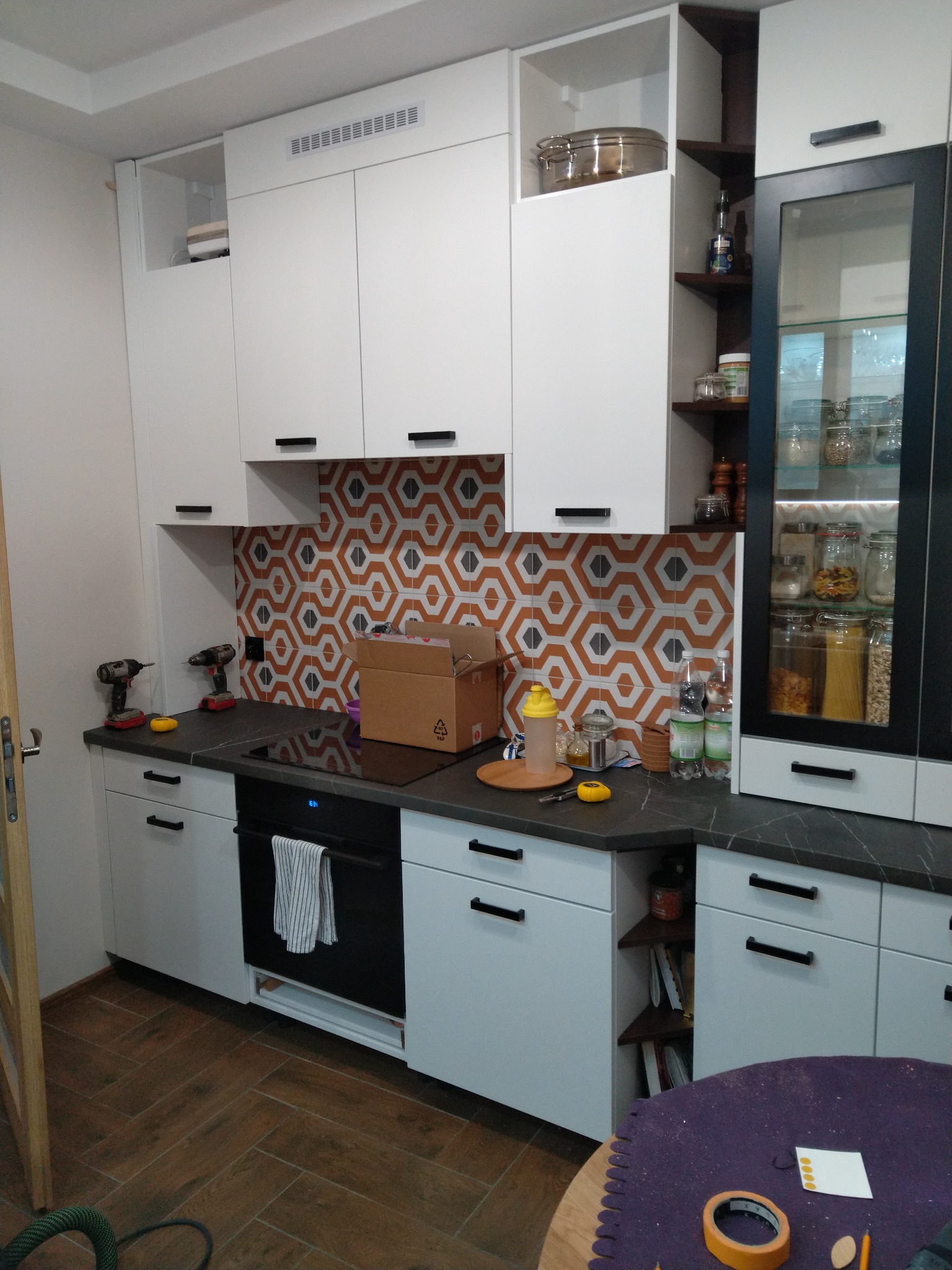 Montaż białej zabudowy kuchennej z czarnymi uchwytami, widoczne narzędzia, kartonowe pudło i płytki na ścianie w geometryczny wzór.