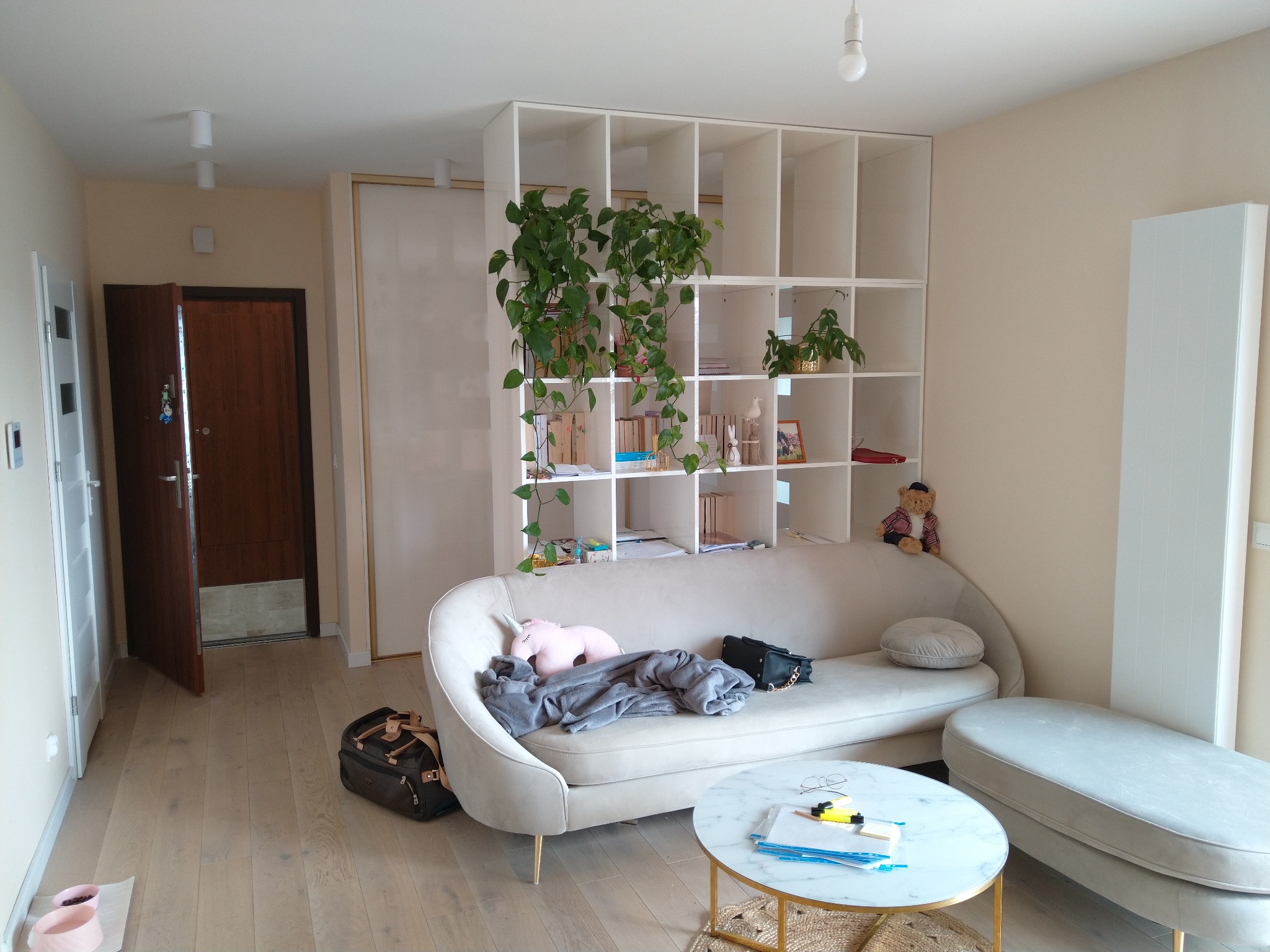 Biały regał z półkami, na których stoją rośliny i dekoracje, ustawiony za beżową sofą w jasnym salonie z drewnianą podłogą.