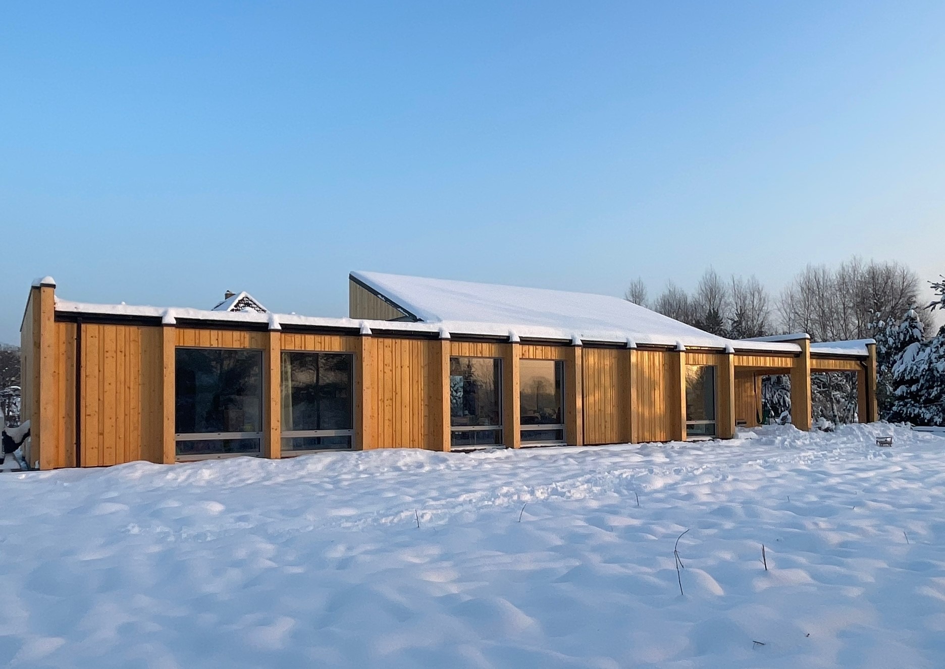 Nowoczesny dom jednorodzinny z drewnianą elewacją i dużymi oknami, częściowo zasypany śniegiem, sfotografowany w zimowy dzień przy błękitnym niebie.