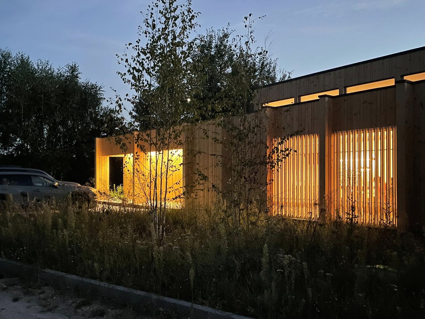 Nowoczesny budynek usługowy z drewnianą elewacją i pionowymi żaluzjami okiennymi, oświetlony ciepłym światłem wieczornym, z samochodem zaparkowanym obok i wysoką trawą na pierwszym planie.