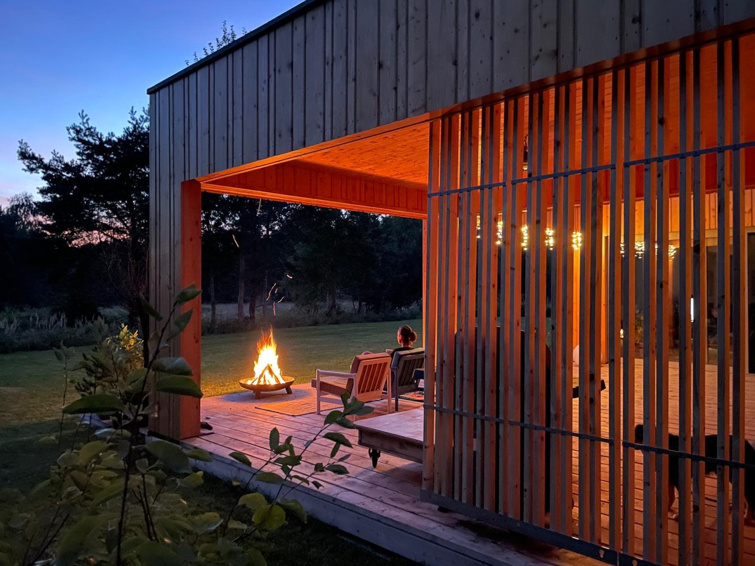 Nowoczesny drewniany dom z tarasem oświetlonym wieczornym światłem, z paleniskiem na zewnątrz i osobą siedzącą w fotelu, widok przez ażurową ścianę.