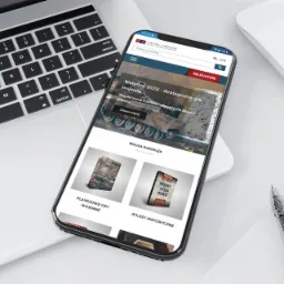 Smartfon wyświetla mobilną wersję sklepu internetowego z grami strategicznymi i atlasami historycznymi, leżący obok laptopa, myszki i notatnika z długopisem.