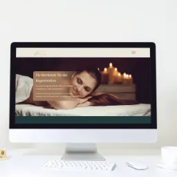 Ekran komputera wyświetla stronę internetową z motywem spa, przedstawiającą kobietę relaksującą się podczas zabiegu, w tle świece, na biurku obok komputera wazon z kwiatami, kalendarz i filiżanka.