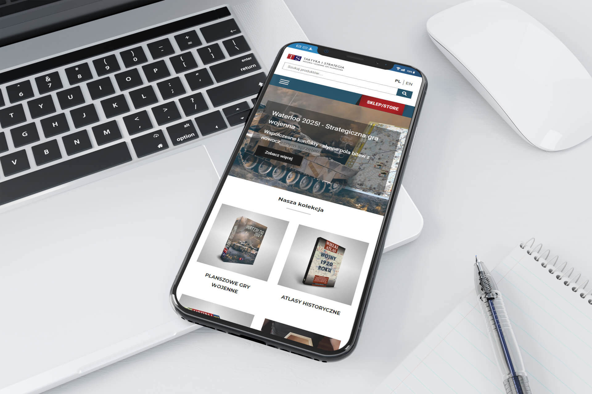 Smartfon wyświetla mobilną wersję sklepu internetowego z grami strategicznymi i atlasami historycznymi, leżący obok laptopa, myszki i notatnika z długopisem.