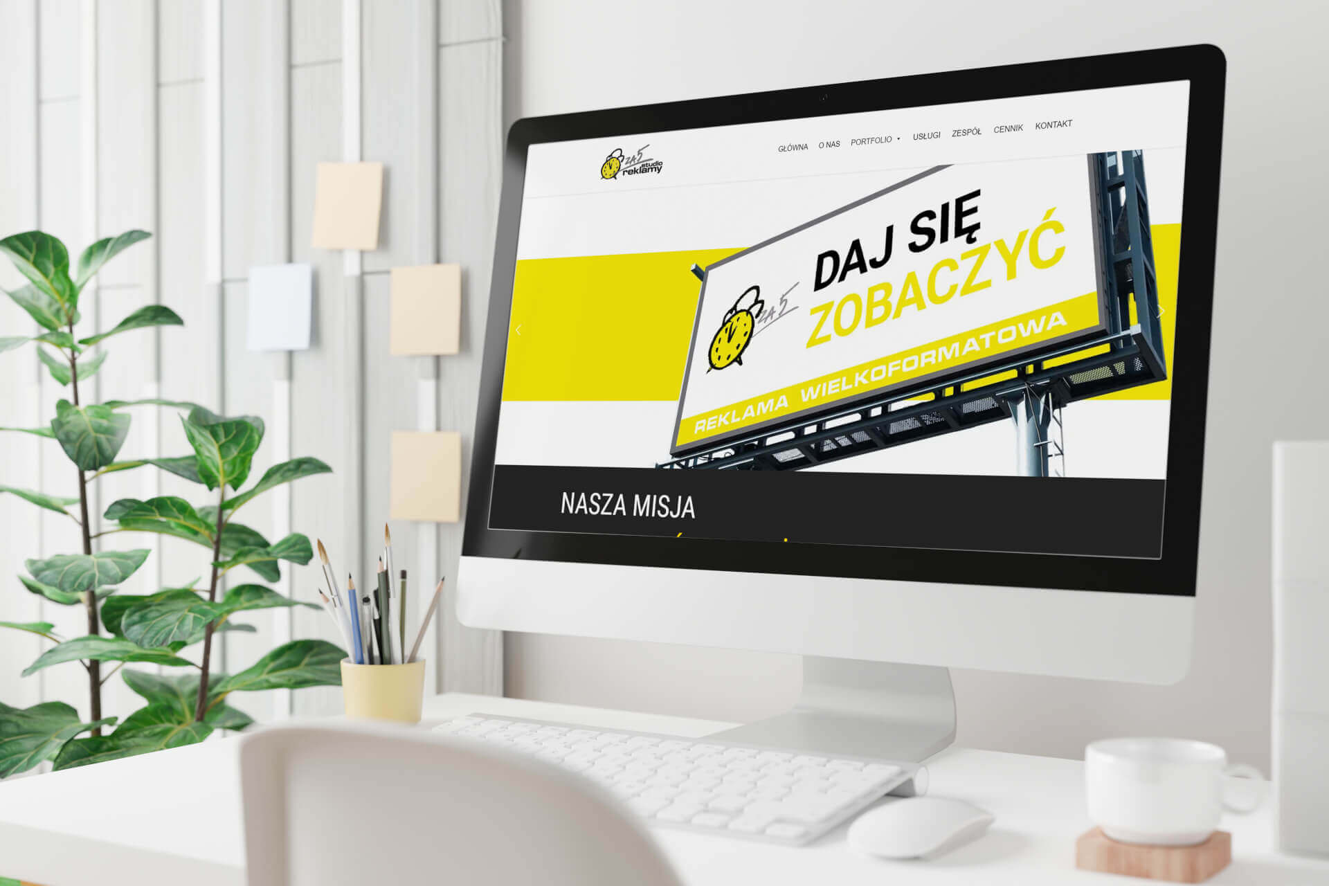 Ekran komputera stacjonarnego wyświetlający stronę internetową z billboardem reklamowym, obok roślina doniczkowa i przybory do pisania, kubek z kawą na biurku.