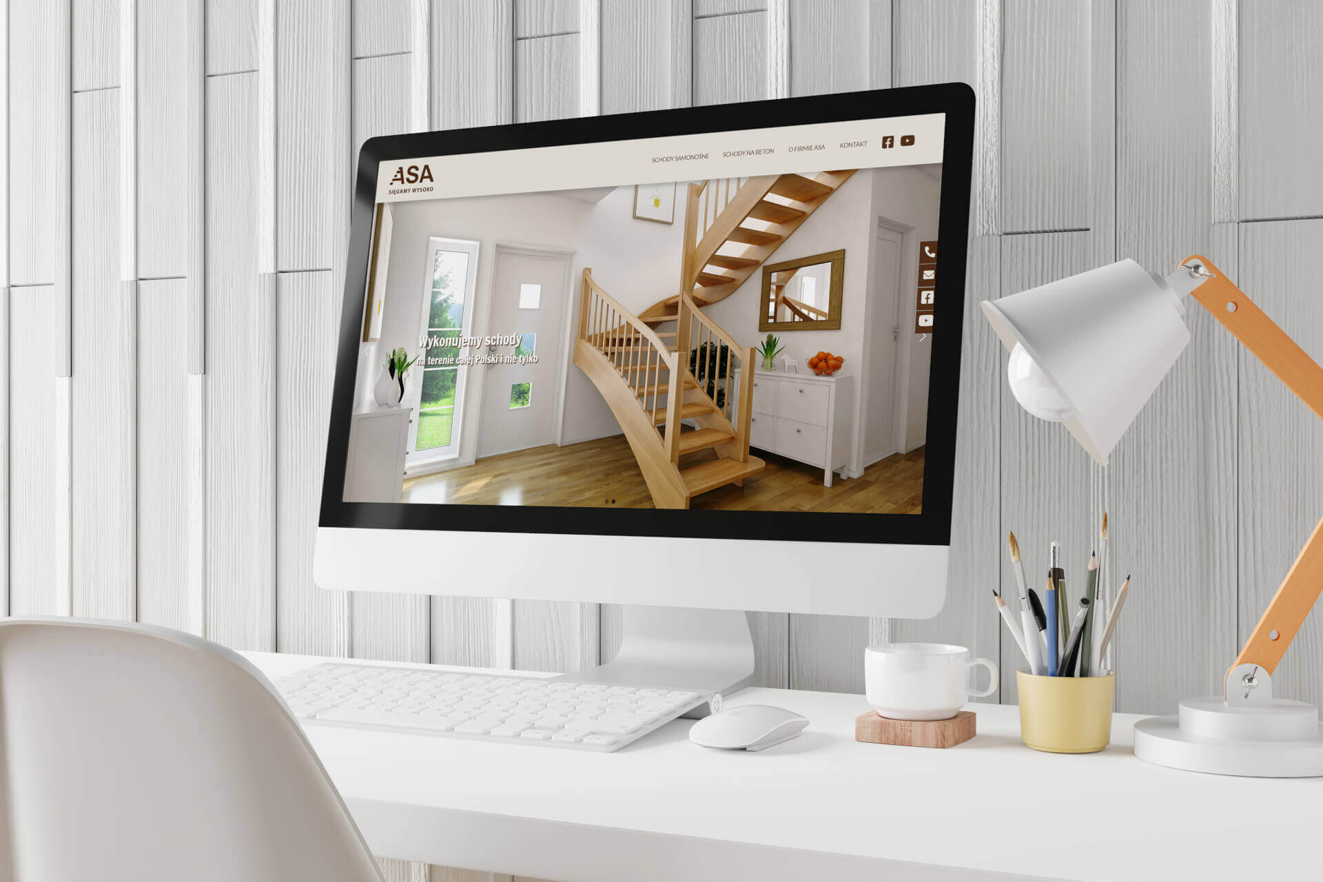Monitor wyświetla stronę internetową firmy ASA prezentującą drewniane schody w jasnym wnętrzu, widoczne biurko z lampką, kubkiem i przyborami do pisania.