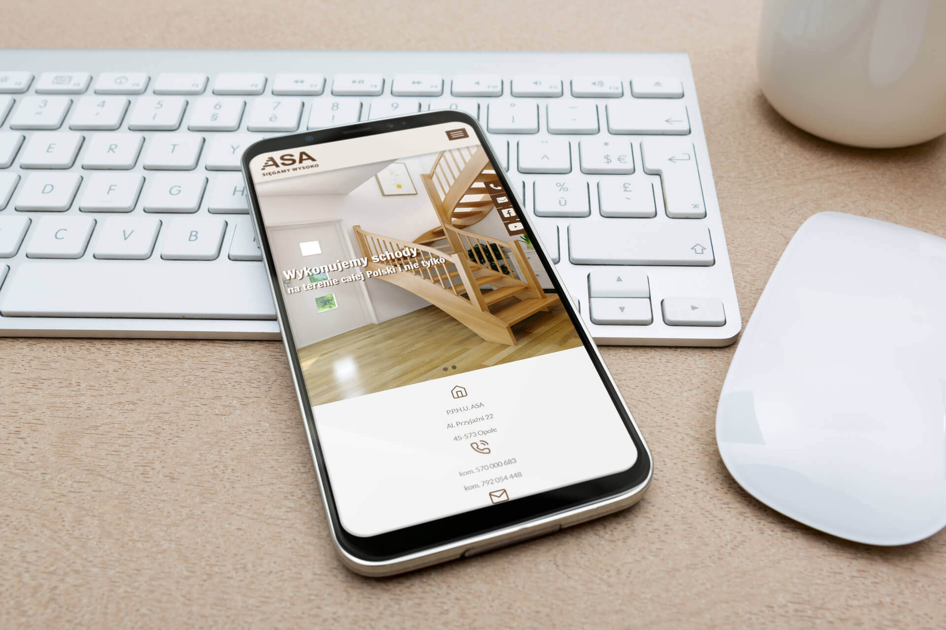 Smartfon wyświetlający stronę internetową firmy ASA z wizualizacją drewnianych schodów, obok biała klawiatura i myszka na drewnianym biurku.