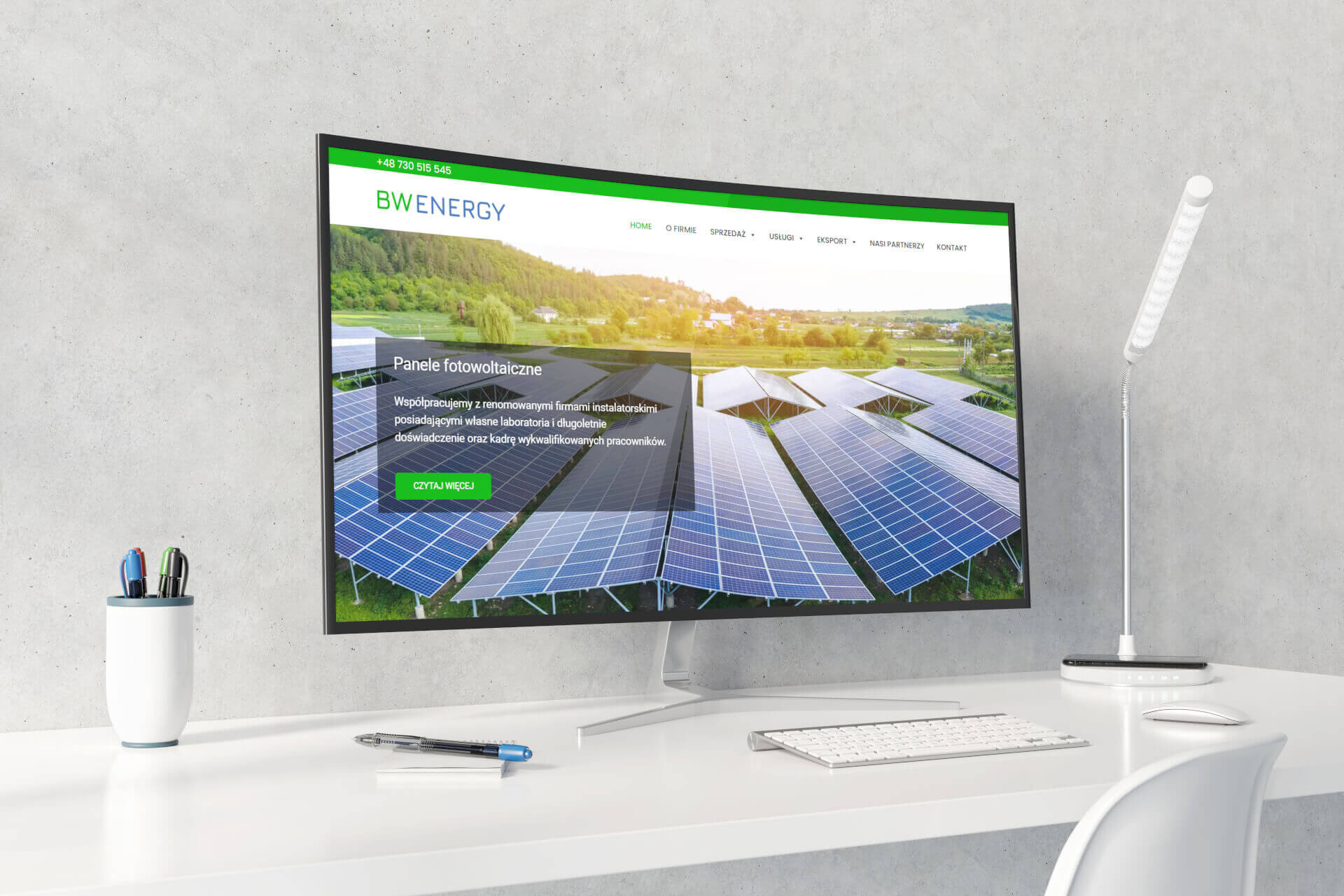 Monitor wyświetla stronę internetową firmy BW Energy z panelami fotowoltaicznymi w tle, obok lampka biurkowa i przybory do pisania na białym biurku.