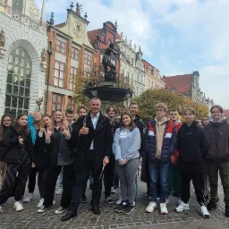 Grupa młodzieży z przewodnikiem pozuje na tle zabytkowej fontanny Neptuna i kolorowych kamienic w Gdańsku.