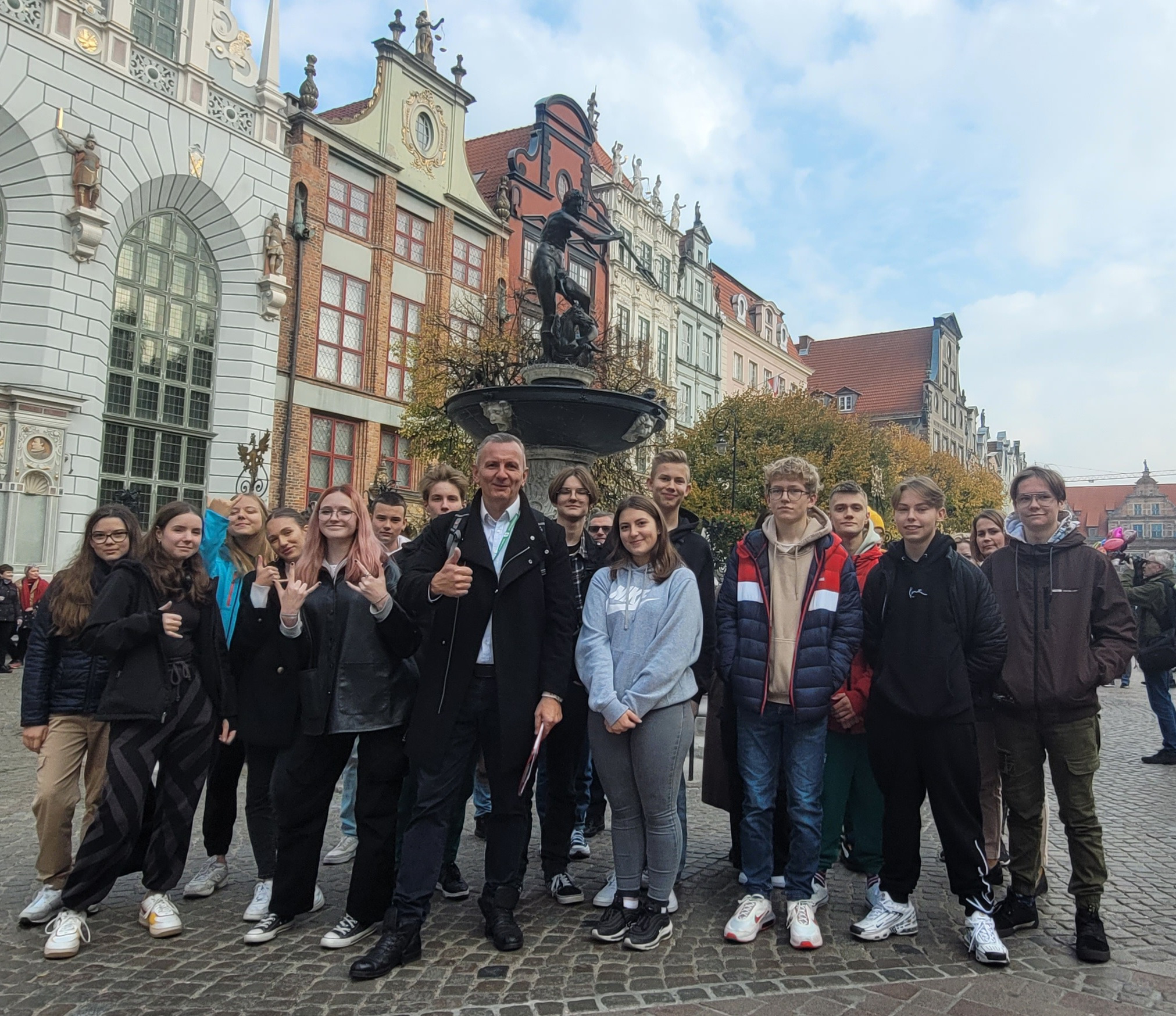 Grupa młodzieży z przewodnikiem pozuje na tle zabytkowej fontanny Neptuna i kolorowych kamienic w Gdańsku.