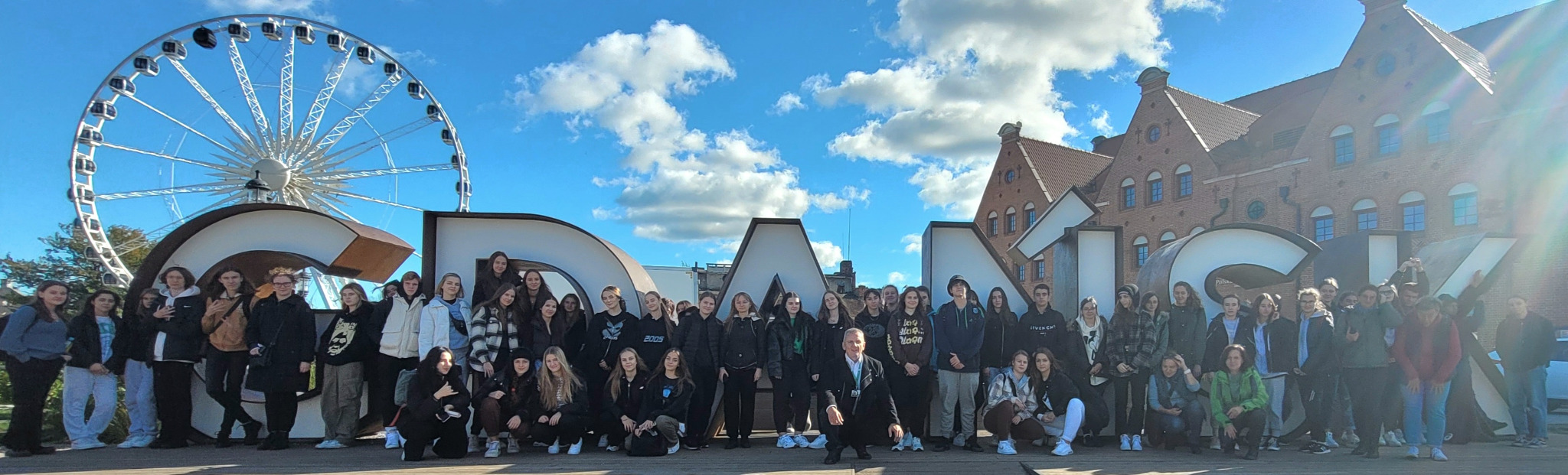 Grupa uczniów pozuje na tle napisu Gdańsk i diabelskiego młyna w słoneczny dzień.