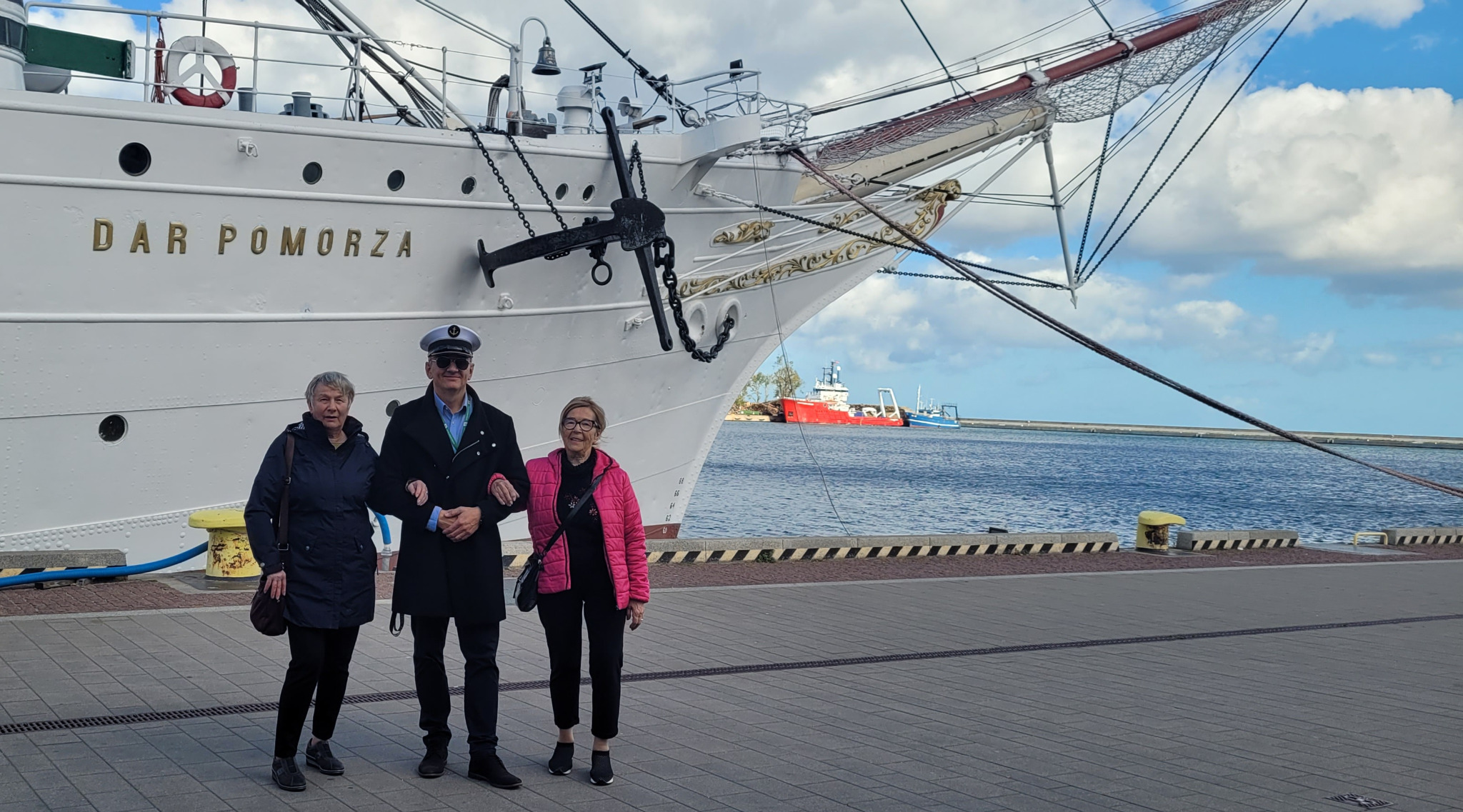 Trzy osoby pozują na tle statku Dar Pomorza w Gdyni, kapitan w mundurze i okularych słonecznych trzyma pod ramię dwie kobiety, w tle widoczne morze i inne statki.