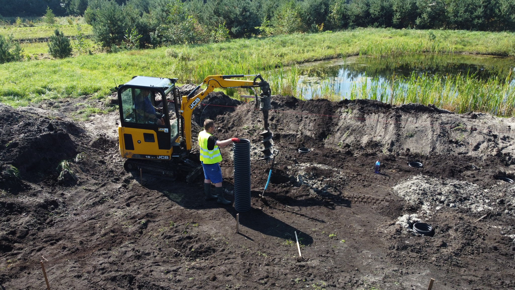 Żółta minikoparka JCB z wiertnicą wierci otwór w ziemi obok czarnej rury drenażowej, w tle zielony teren z oczkiem wodnym.
