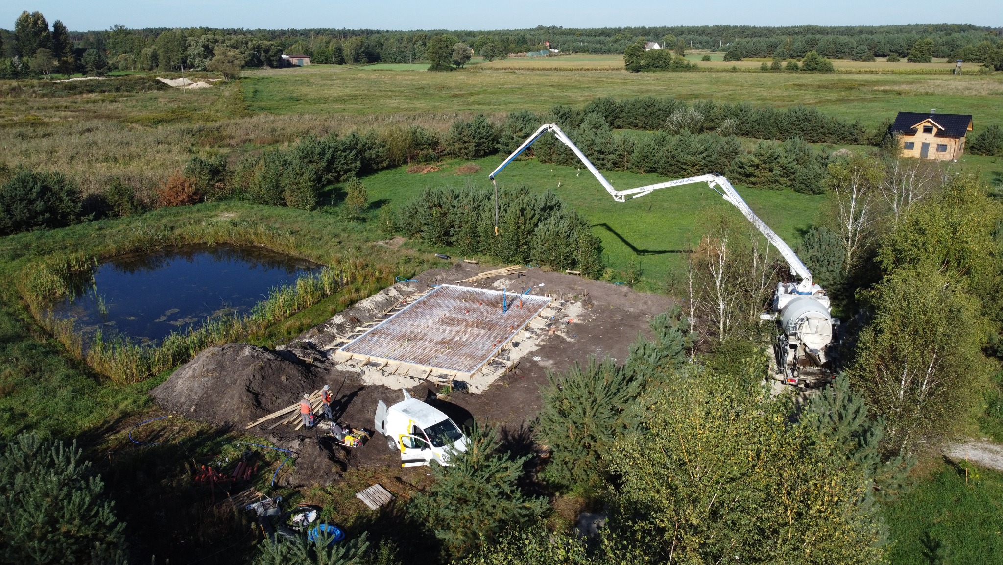 Widok z góry: budowa fundamentów domu jednorodzinnego obok stawu, z użyciem pompy do betonu, widoczna stal zbrojeniowa i szalunki. Dwie osoby w odblaskowych kamizelkach nadzorują pracę.