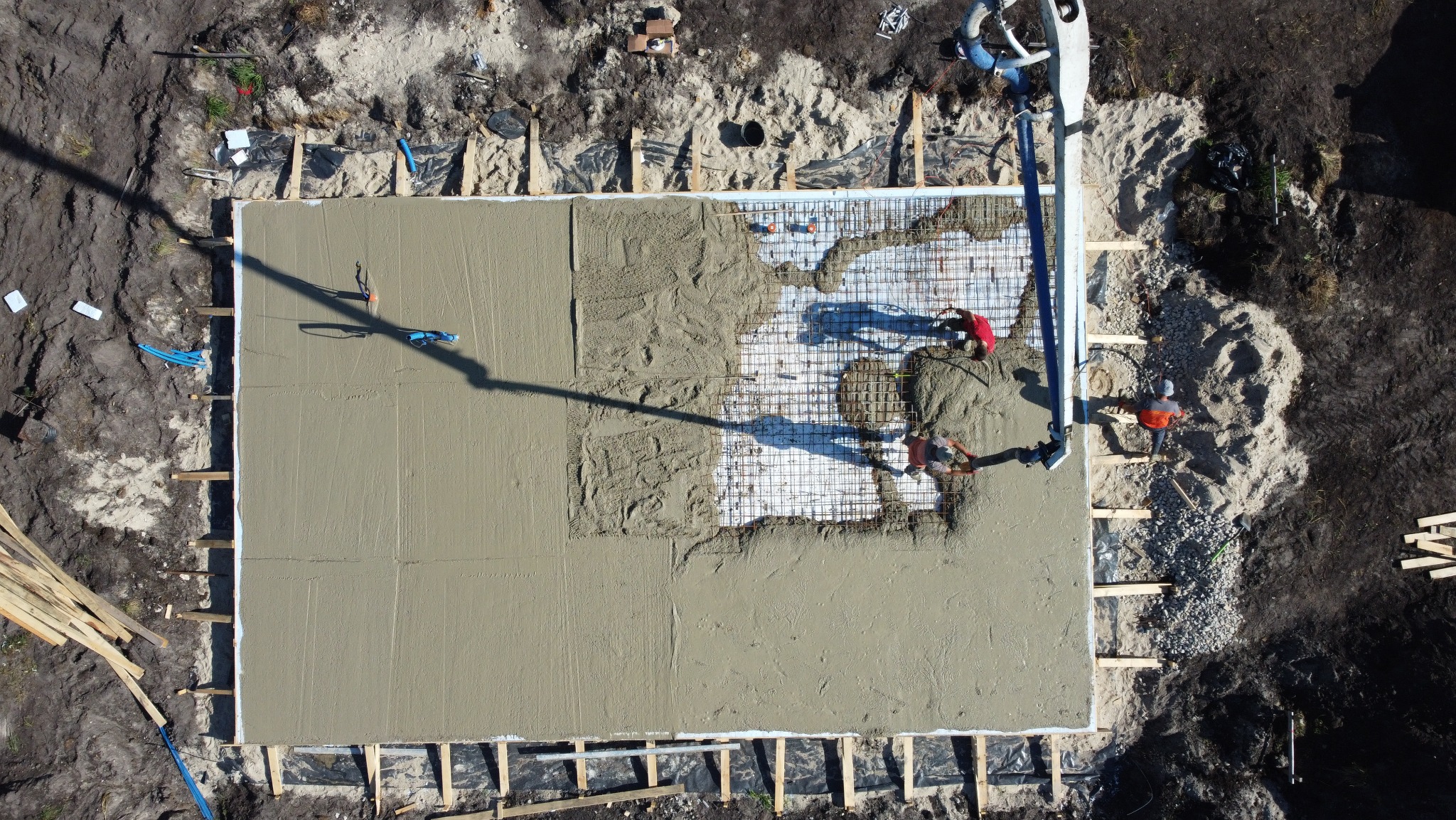 Zalewanie betonem zbrojonej płyty fundamentowej z użyciem pompy do betonu, widok z góry, dwóch pracowników w kaskach i pomarańczowych kamizelkach, częściowo zalany obszar, drewniane szalunki.