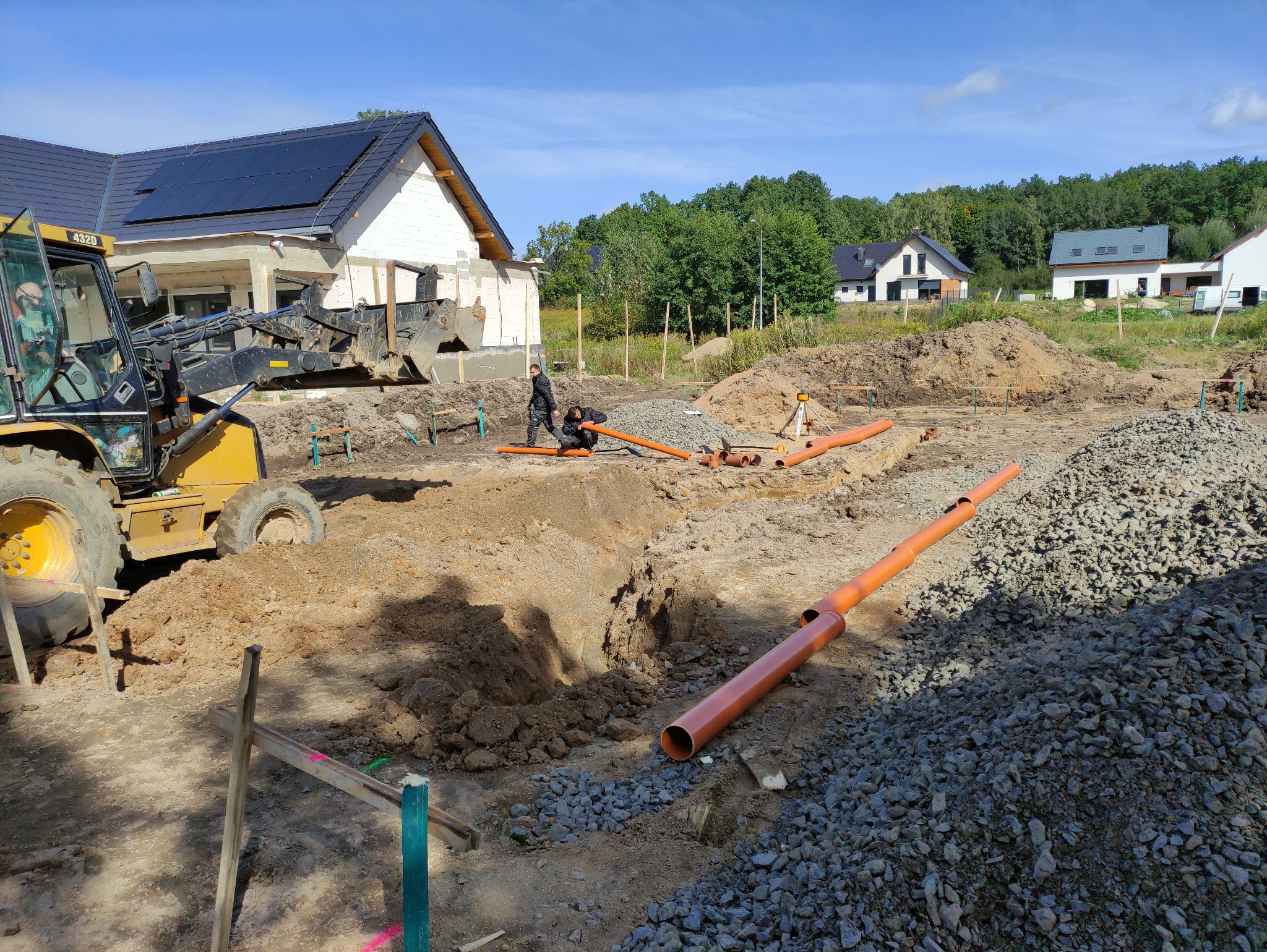 Budowa fundamentów domu jednorodzinnego: widok na wykop pod fundament, ułożone rury kanalizacyjne, koparkę i pracowników przy montażu instalacji, w tle dom w stanie surowym otwartym.