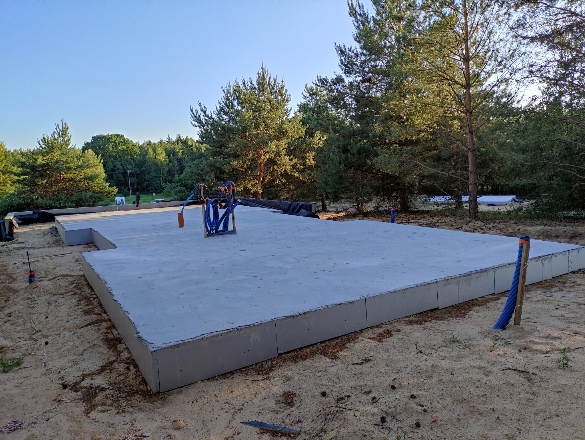 Wylana płyta fundamentowa na piaszczystym terenie z widocznymi przyłączami wodno-kanalizacyjnymi, w tle las iglasty pod błękitnym niebem.