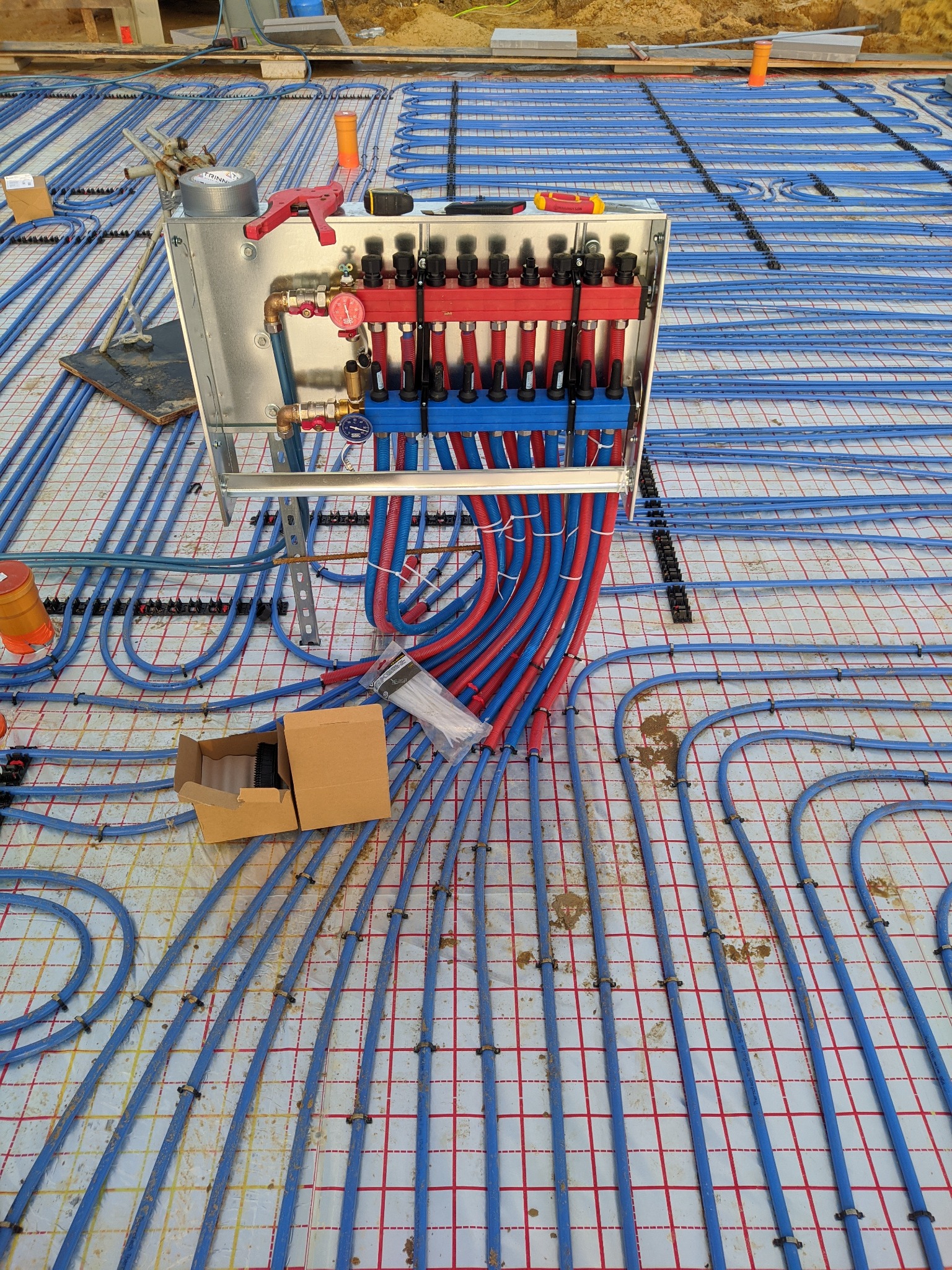 Rozdzielacz ogrzewania podłogowego z czerwonymi i niebieskimi rurami ułożonymi na folii z siatką, widoczne narzędzia i kartonowe pudełko.