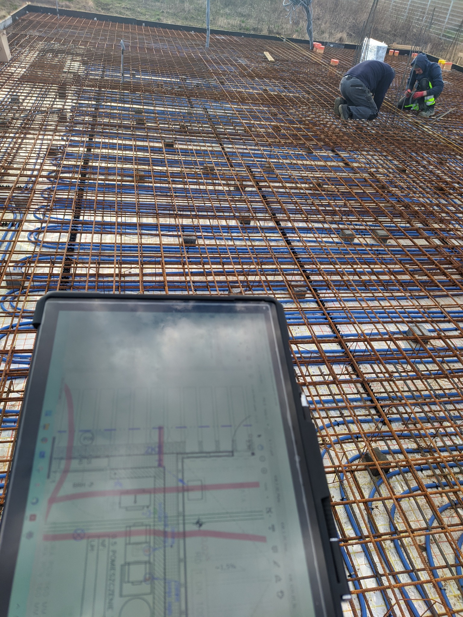 Zbrojenie płyty fundamentowej z ogrzewaniem podłogowym, widok z góry z tabletem prezentującym plan budynku w pierwszym planie, dwóch pracowników w tle.