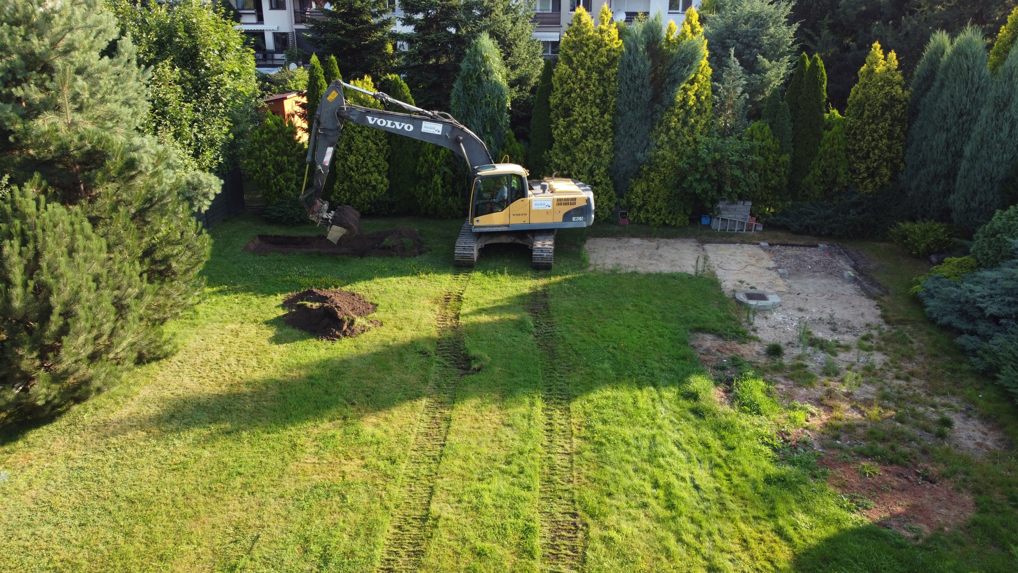 Żółty koparka Volvo EC210C wykopuje dół pod fundament na trawiastym terenie z widocznymi śladami gąsienic, w tle zabudowa jednorodzinna i drzewa iglaste.