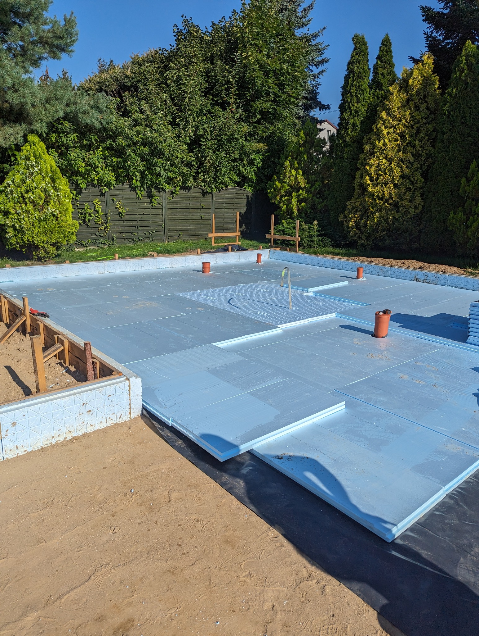 Izolacja termiczna z płyt styropianowych na przygotowanym fundamencie, widoczne rury instalacyjne i szalunek drewniany.