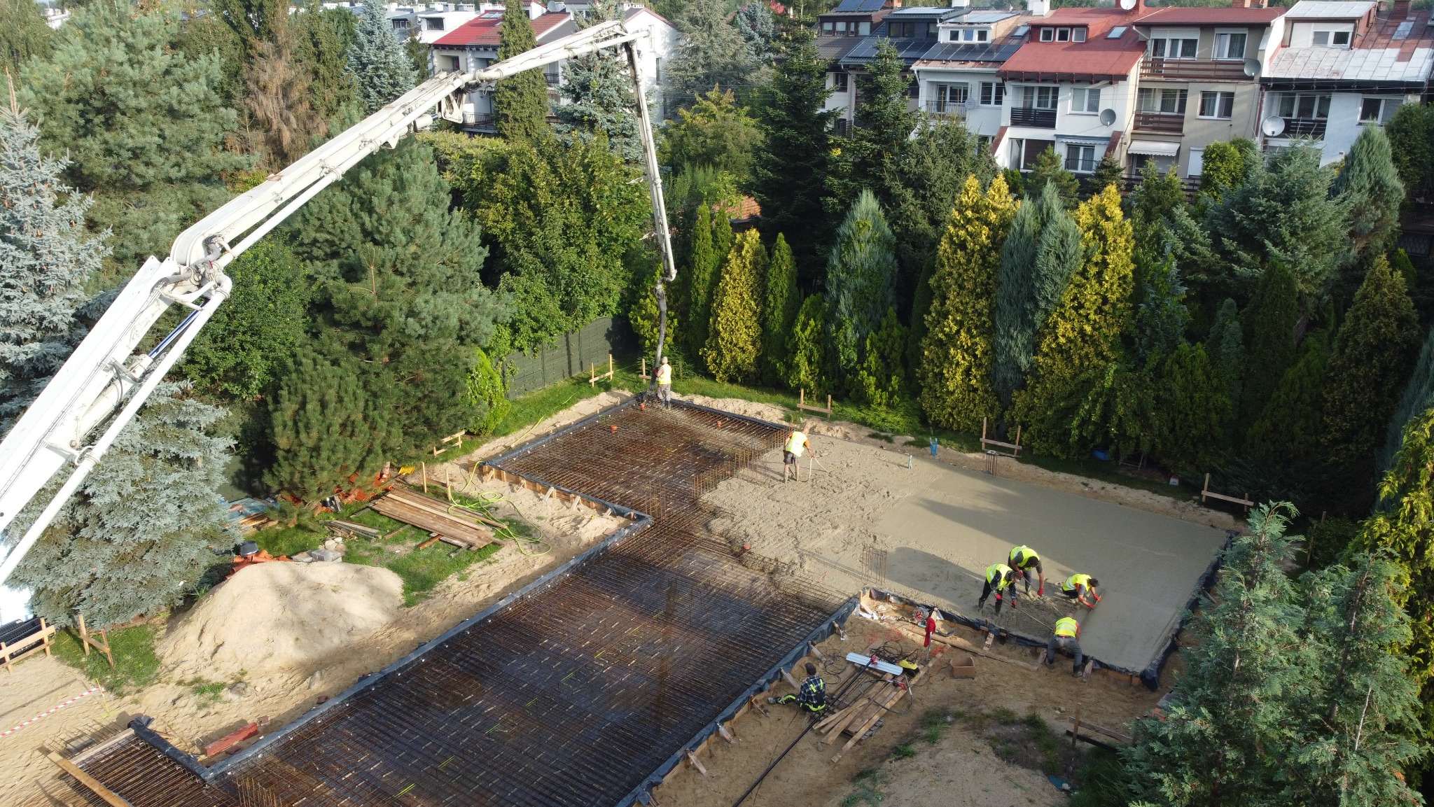 Zalewanie betonem zbrojonej płyty fundamentowej domu jednorodzinnego z użyciem pompy do betonu, widok z góry na plac budowy otoczony zielenią.