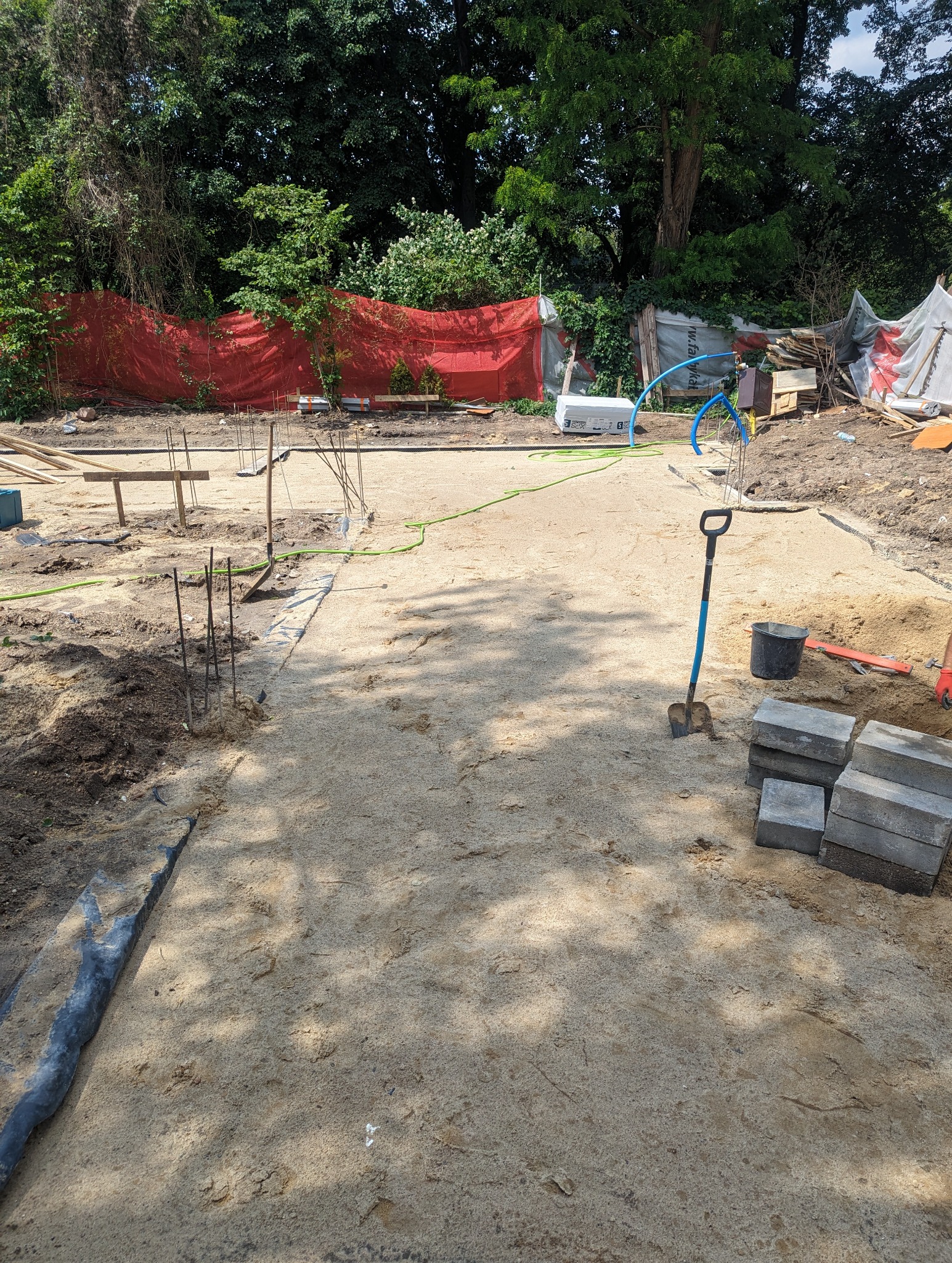 Przygotowany teren pod wylewkę, widoczne wystające pręty zbrojeniowe, szpadel z niebieską rączką, ułożone kostki brukowe, zielony wąż ogrodowy i czerwona siatka w tle.