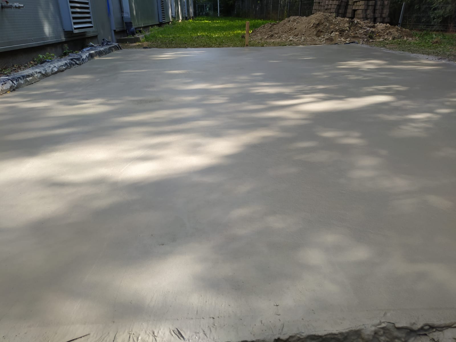 Świeżo wylany betonowy fundament z widocznymi cieniami drzew, zabezpieczony folią budowlaną przy krawędziach, obok budynku z klimatyzacją i stertą cegieł w tle.