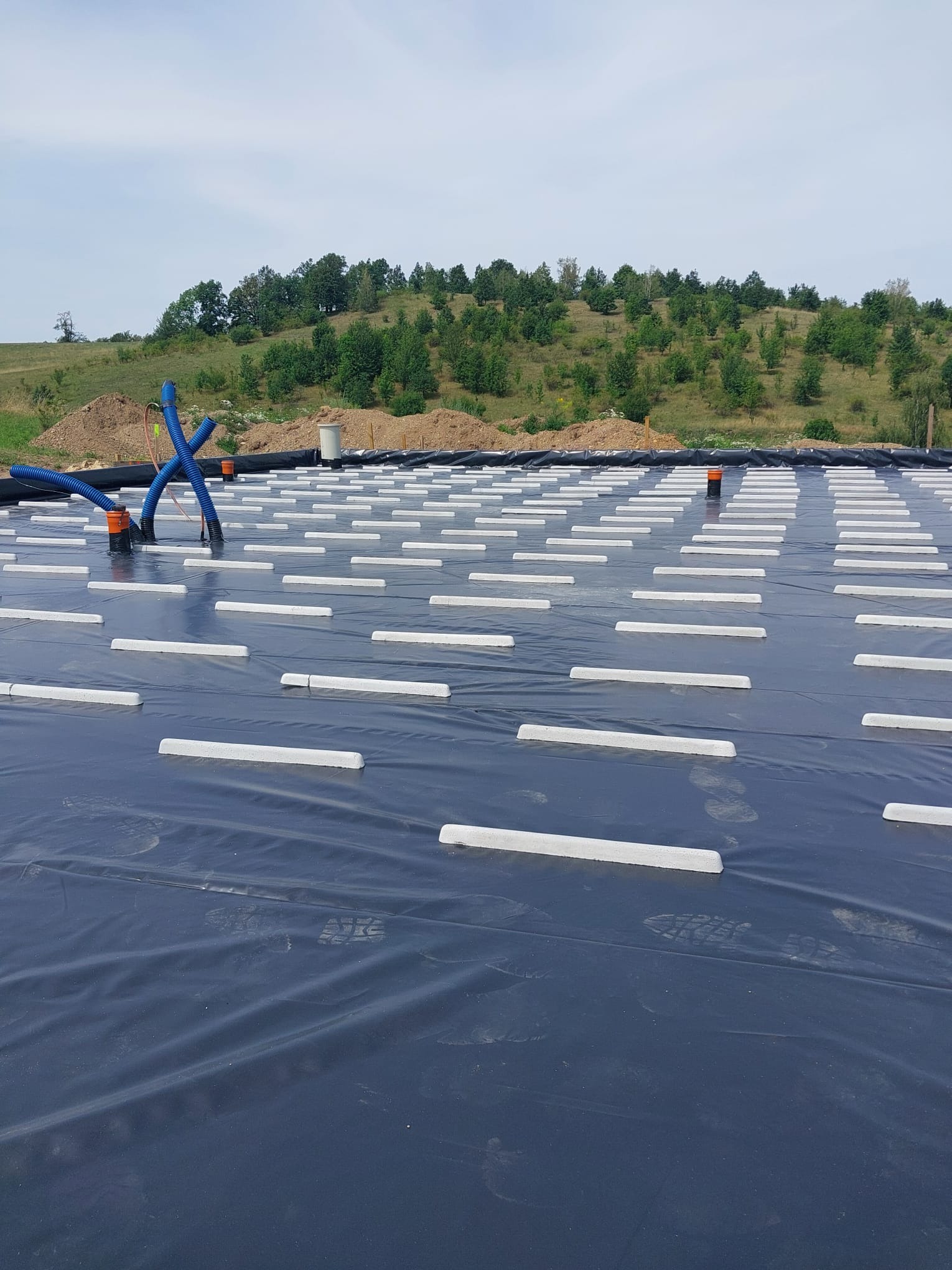 Przygotowanie podłoża pod płytę fundamentową z izolacją poziomą, widoczne białe dystanse na czarnej folii, z niebieskimi rurami wentylacyjnymi. W tle pagórkowaty teren z zielenią.