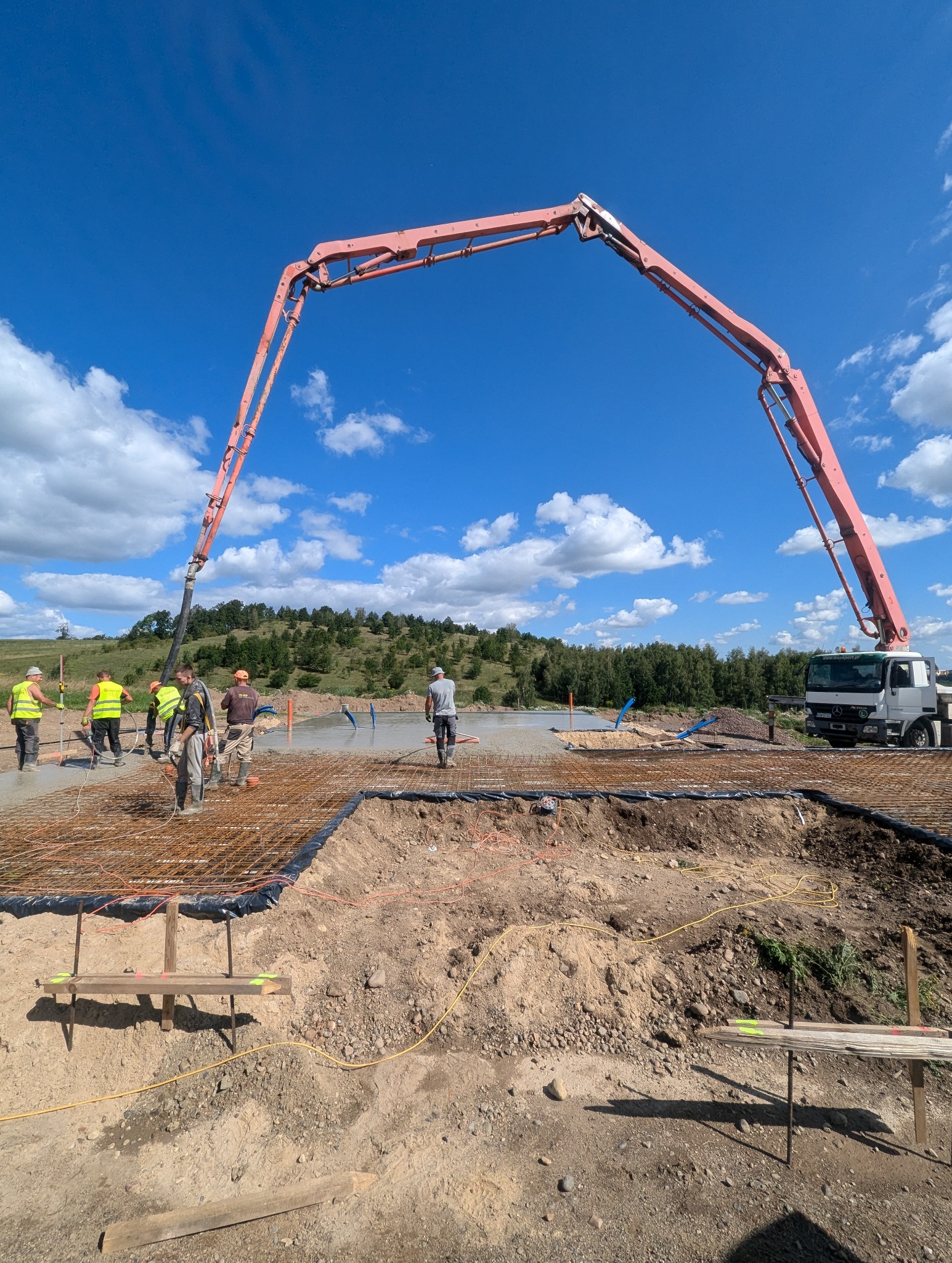 Wylewanie betonu na przygotowane zbrojenie fundamentów za pomocą pompy do betonu; widoczni pracownicy w kamizelkach odblaskowych i kaskach, błękitne niebo z chmurami w tle.