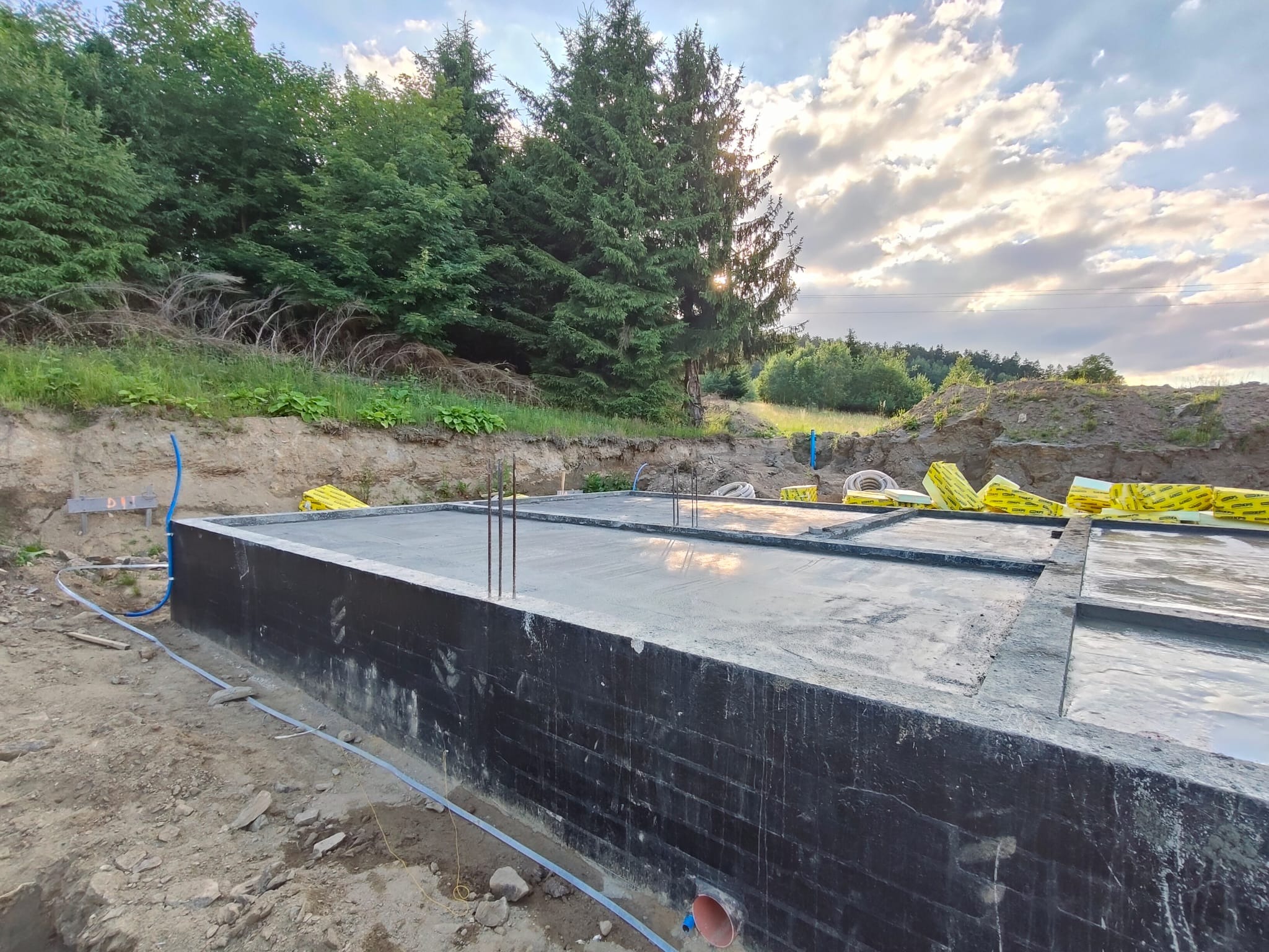 Zalany betonowy fundament z wystającymi prętami zbrojeniowymi, zabezpieczony czarną folią, w wykopie ziemnym z widocznymi rurami instalacyjnymi i zapasem żółtego styropianu.