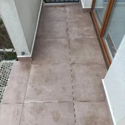 Balkon wyłożony brązowymi płytkami ceramicznymi o matowym wykończeniu, widoczne zabrudzenia i ślady po wodzie, balustrada metalowa w tle, obok okno z drewnianą ramą.