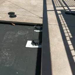 Montaż tarasu wentylowanego: widoczne czarne regulowane wsporniki na membranie hydroizolacyjnej, częściowo ułożone płyty tarasowe i cień metalowej balustrady.